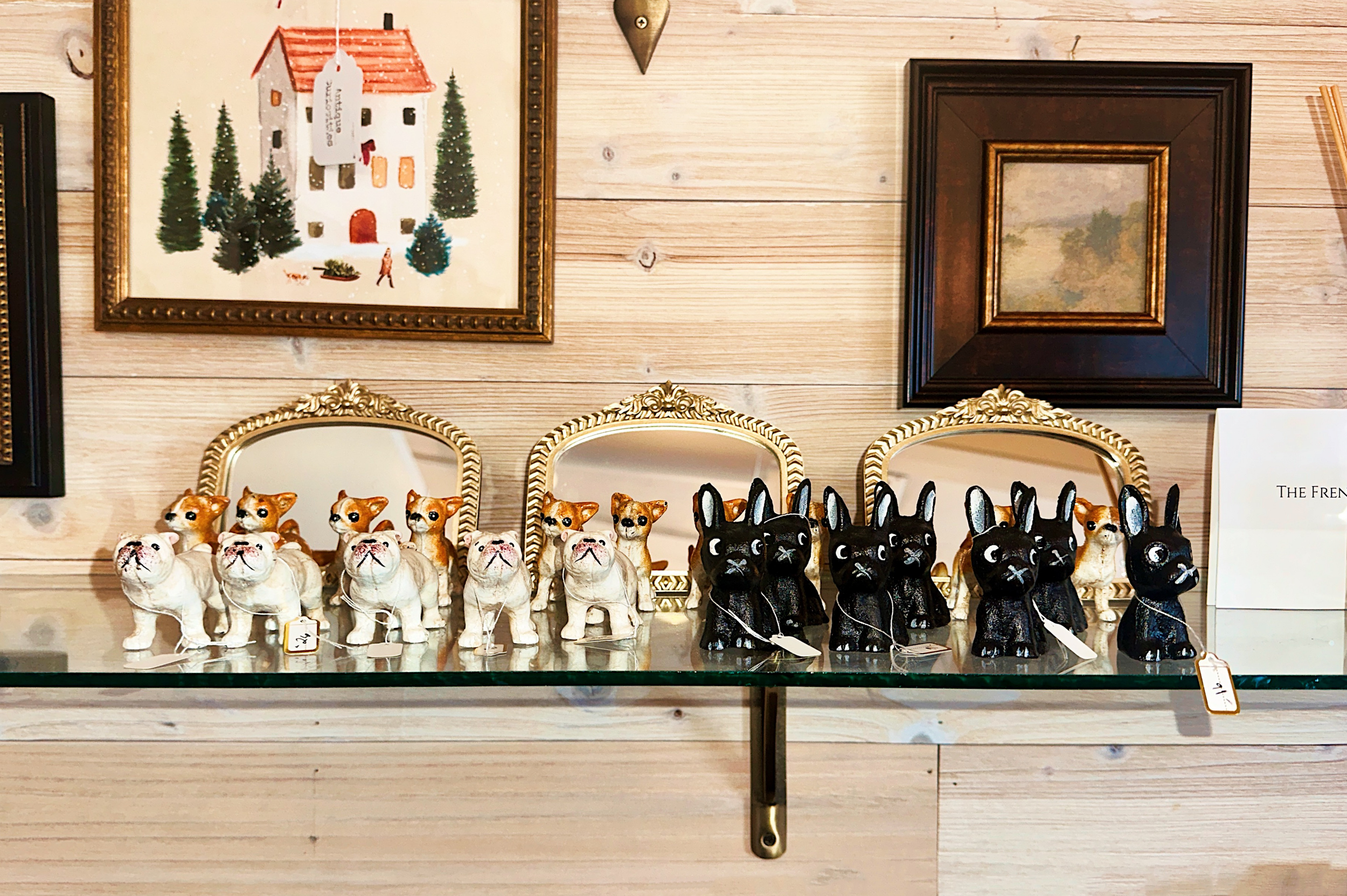 Displays a variety of ceramic figurines of dogs, including bulldogs, grouchy-looking dogs, and black and white dogs, arranged on a glass surface with framed artwork and a wooden wall in the background.