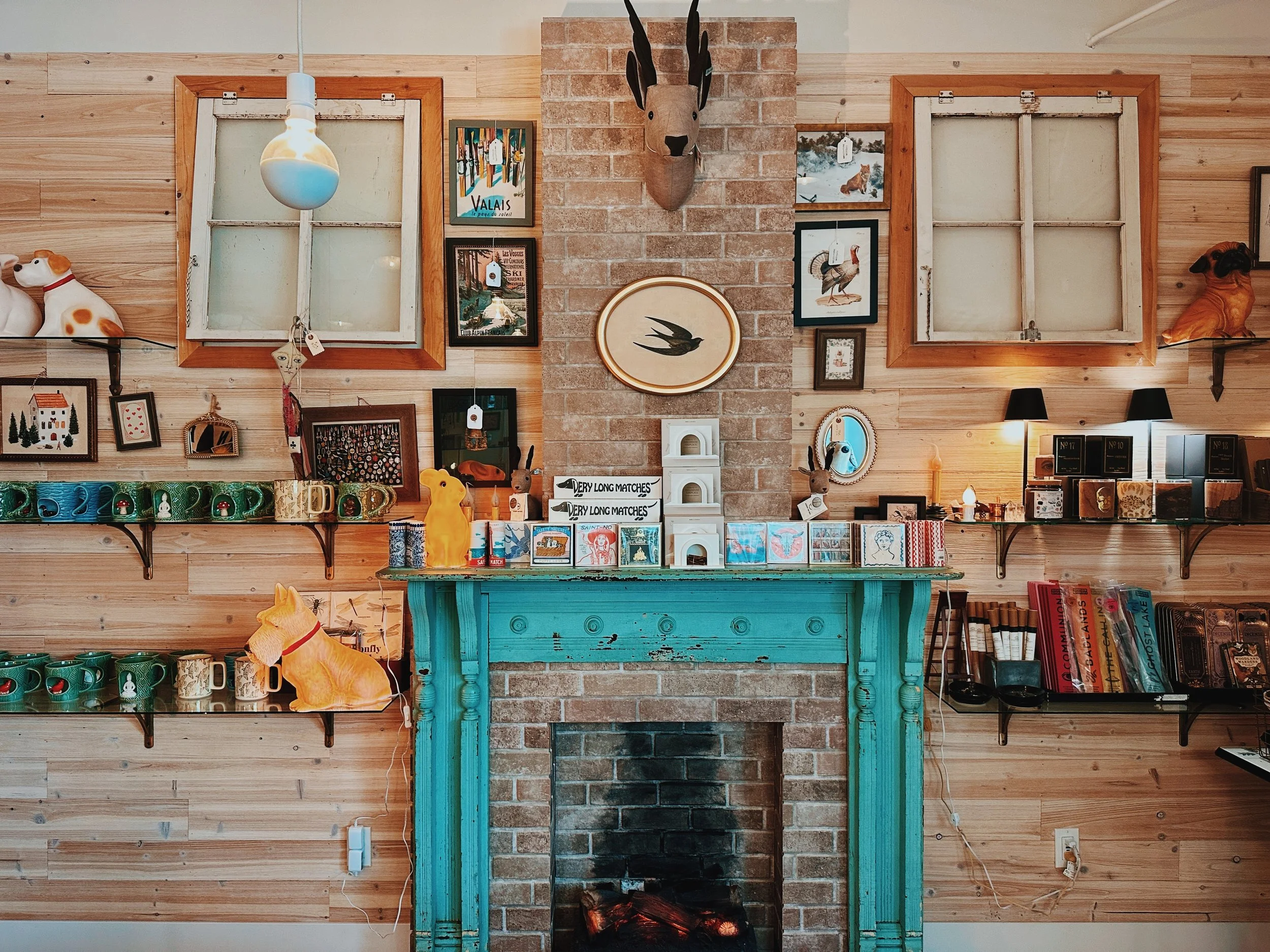 Inside Little Stone. A decorated interior wall with two open windows, framed art, a mounted animal head, plants, and various decorative items on shelves and the mantel.