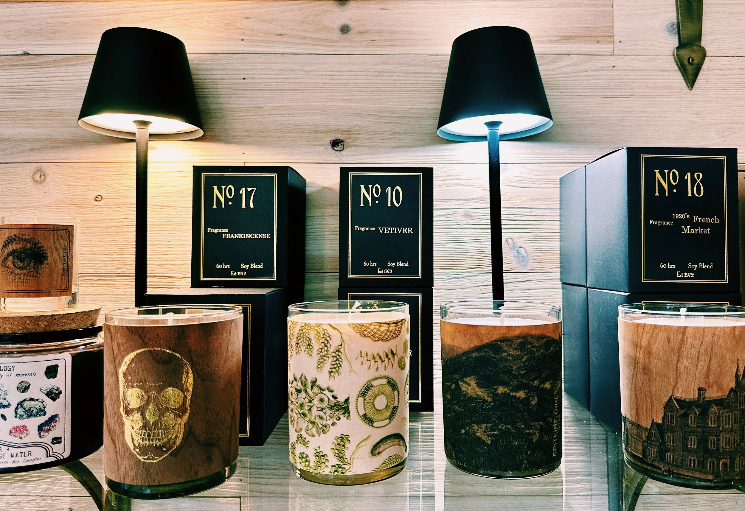 A display of several scented candles with decorative labels and boxes behind them, branded as vintage French fragrances on a wooden surface.