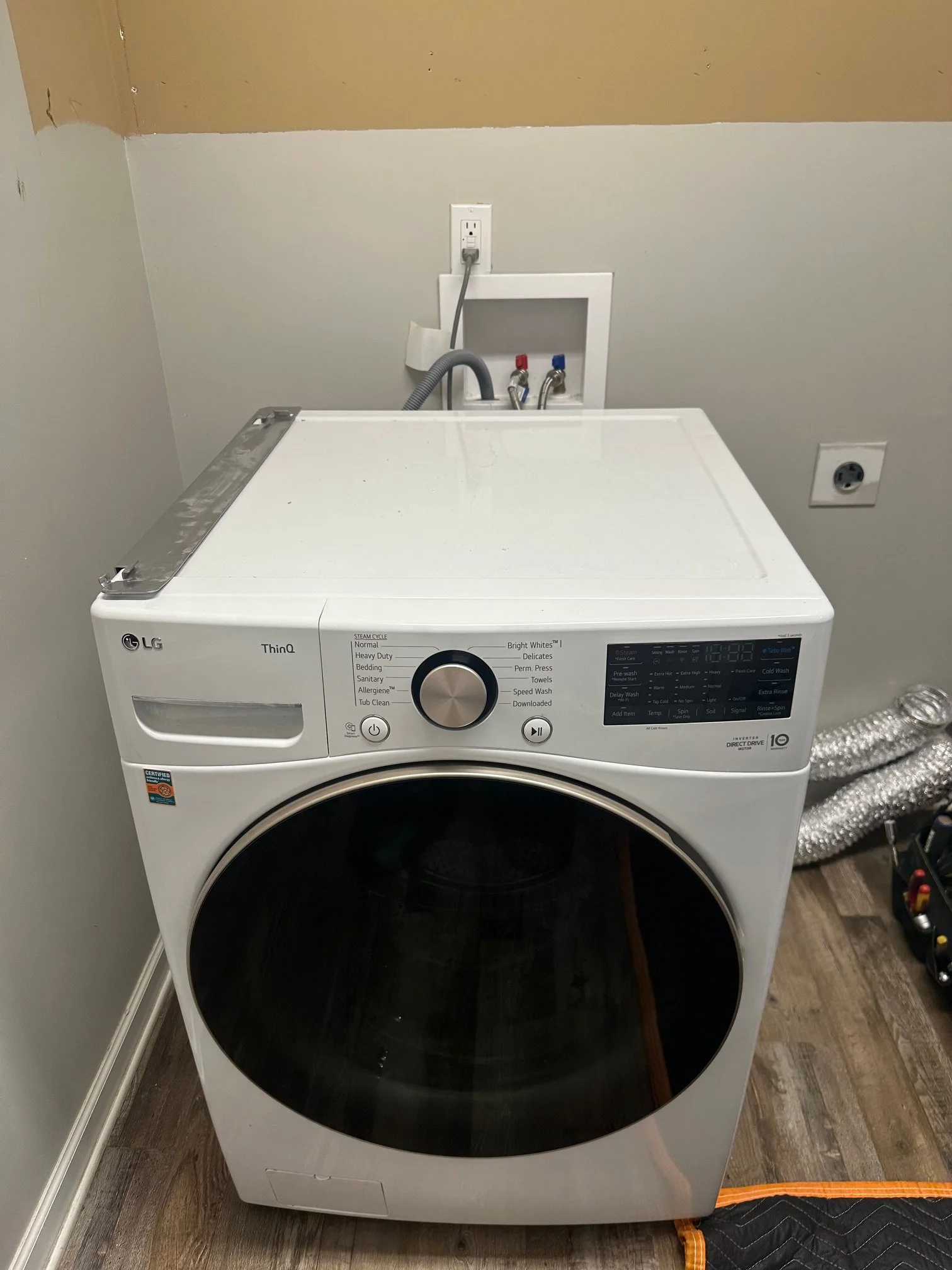 Front view of a white LG ThinQ washing machine in a laundry room, with a black door, control panel, and digital display, positioned against a wall with plumbing connections and electrical outlets behind it.