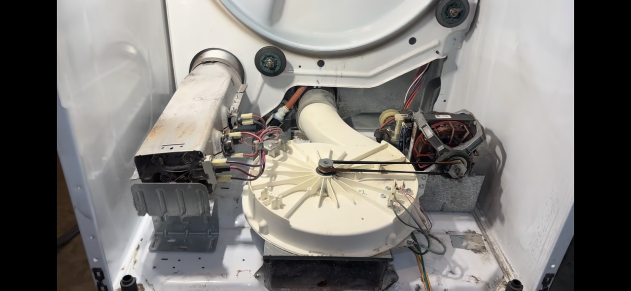 The interior of a washing machine with visible mechanical and electrical components, including a motor, wiring, and pulleys.