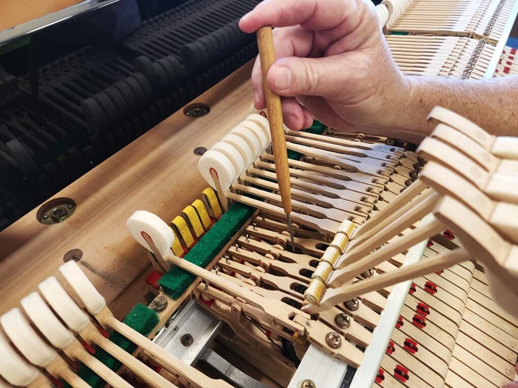 Close-up of a person using a tool to adjust the inside of a piano, showing piano strings and hammers.