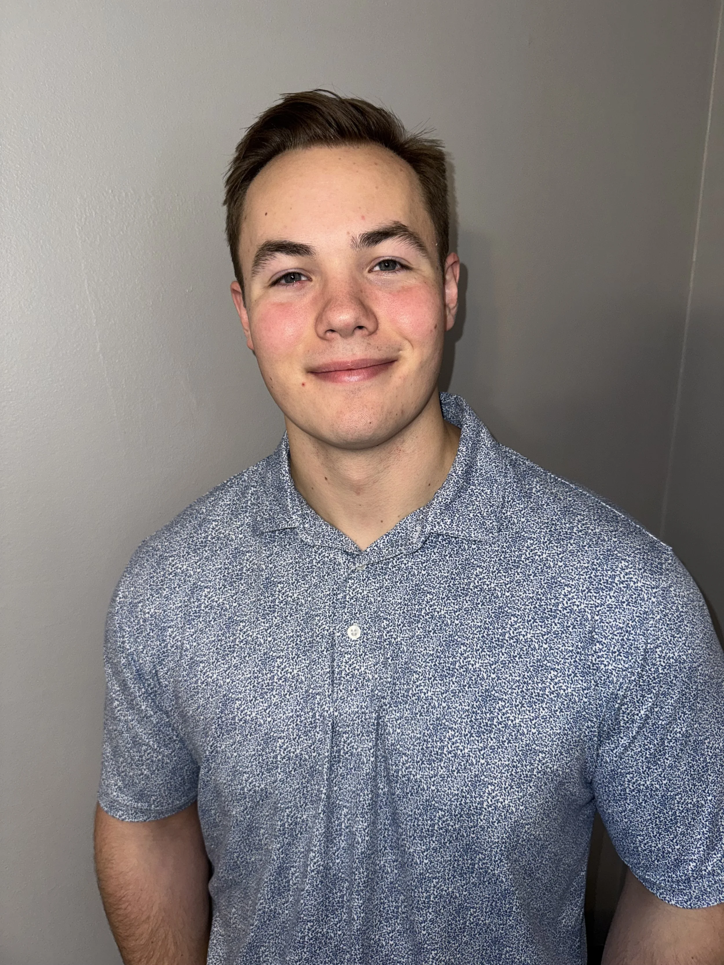 A young man with short brown hair, blue eyes, and fair skin, smiling slightly, wearing a short-sleeved, patterned button-up shirt, standing against a plain gray wall.