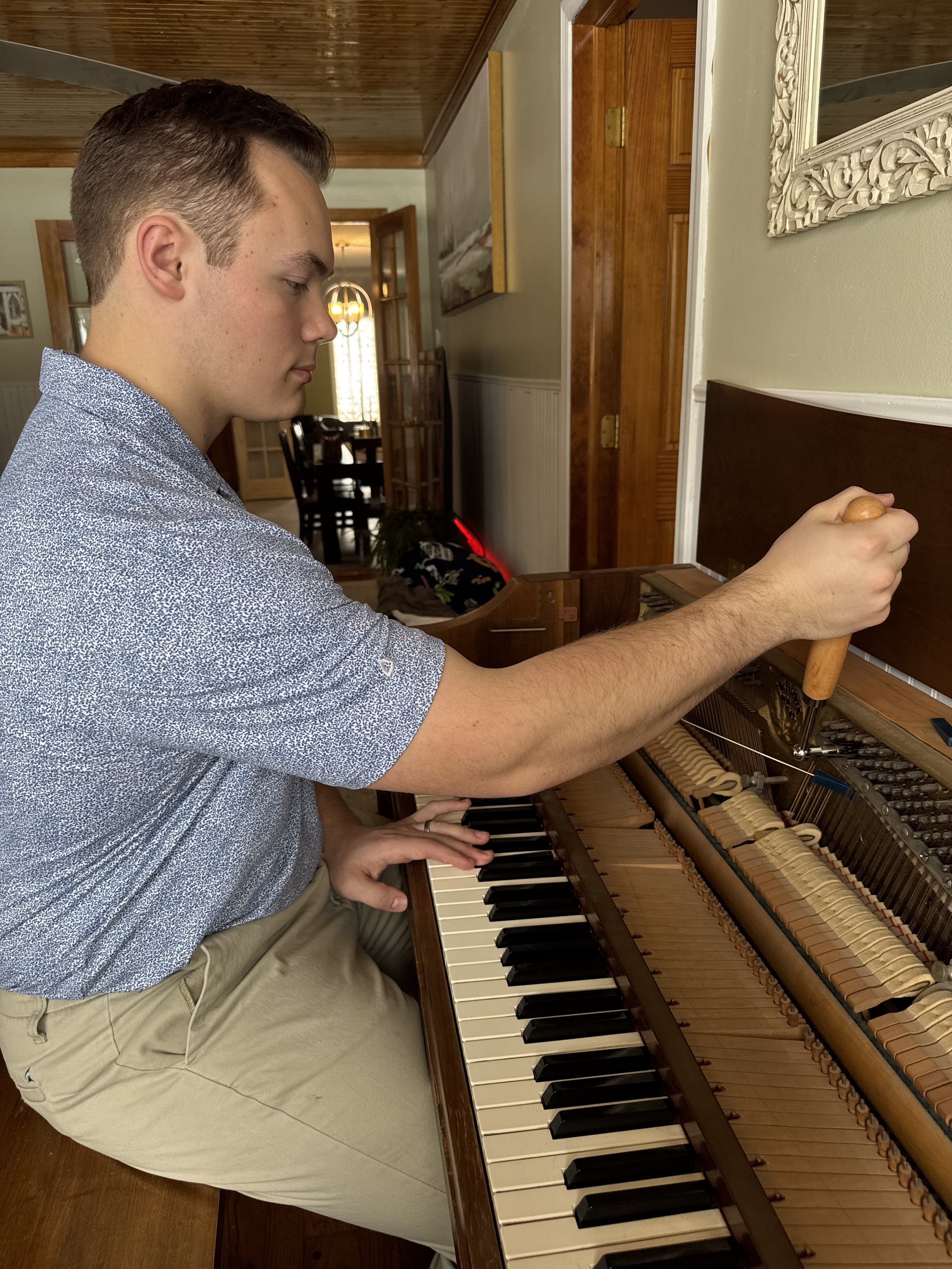 A young man in a blue patterned shirt and beige pants playing a piano in a cozy, well-lit home interior.