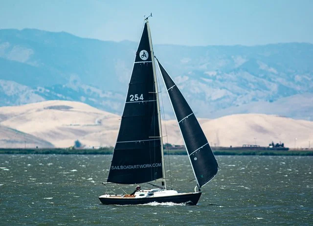 A black and white sailboat with black sails on a body of water, with rolling hills and mountains in the background.