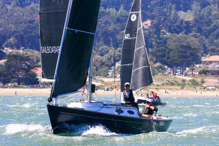 Two people sailing a black and gray sailboat on the water near a sandy beach with houses and trees in the background.