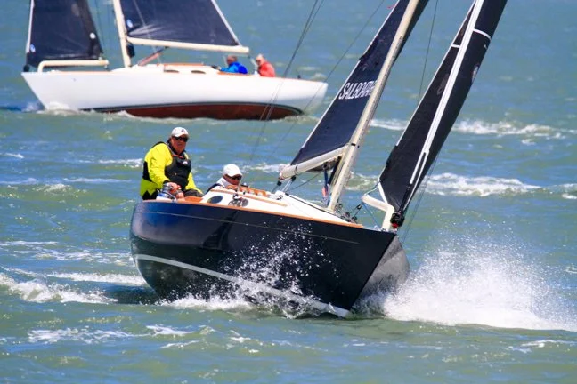 Two sailboats racing on the water, with crew members on board, sails catching the wind, and water splashing against the hulls.