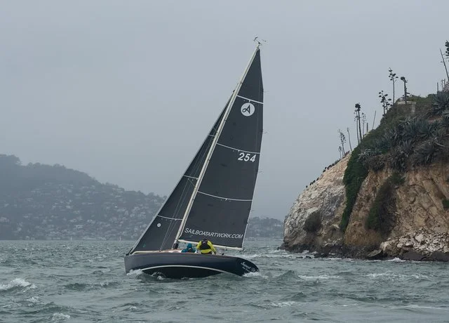 Sailboat racing near rocky coastline with foggy background and a hilly landscape.
