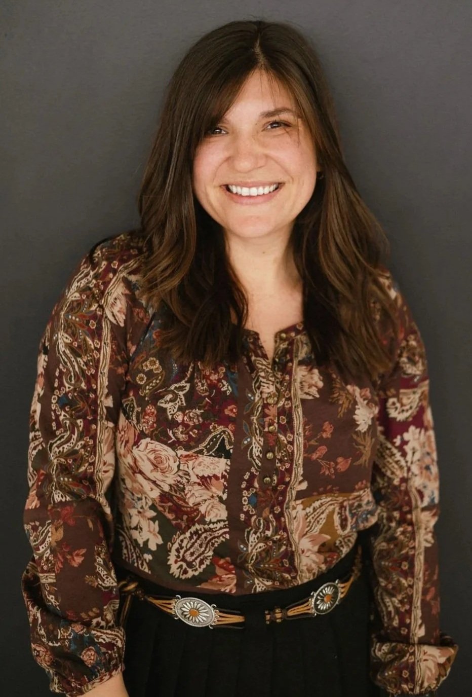 A woman with shoulder-length brown hair, smiling, wearing a long-sleeved, paisley-patterned blouse in shades of brown, red, and cream, with a black belt featuring a silver and floral buckle, standing against a dark gray background.