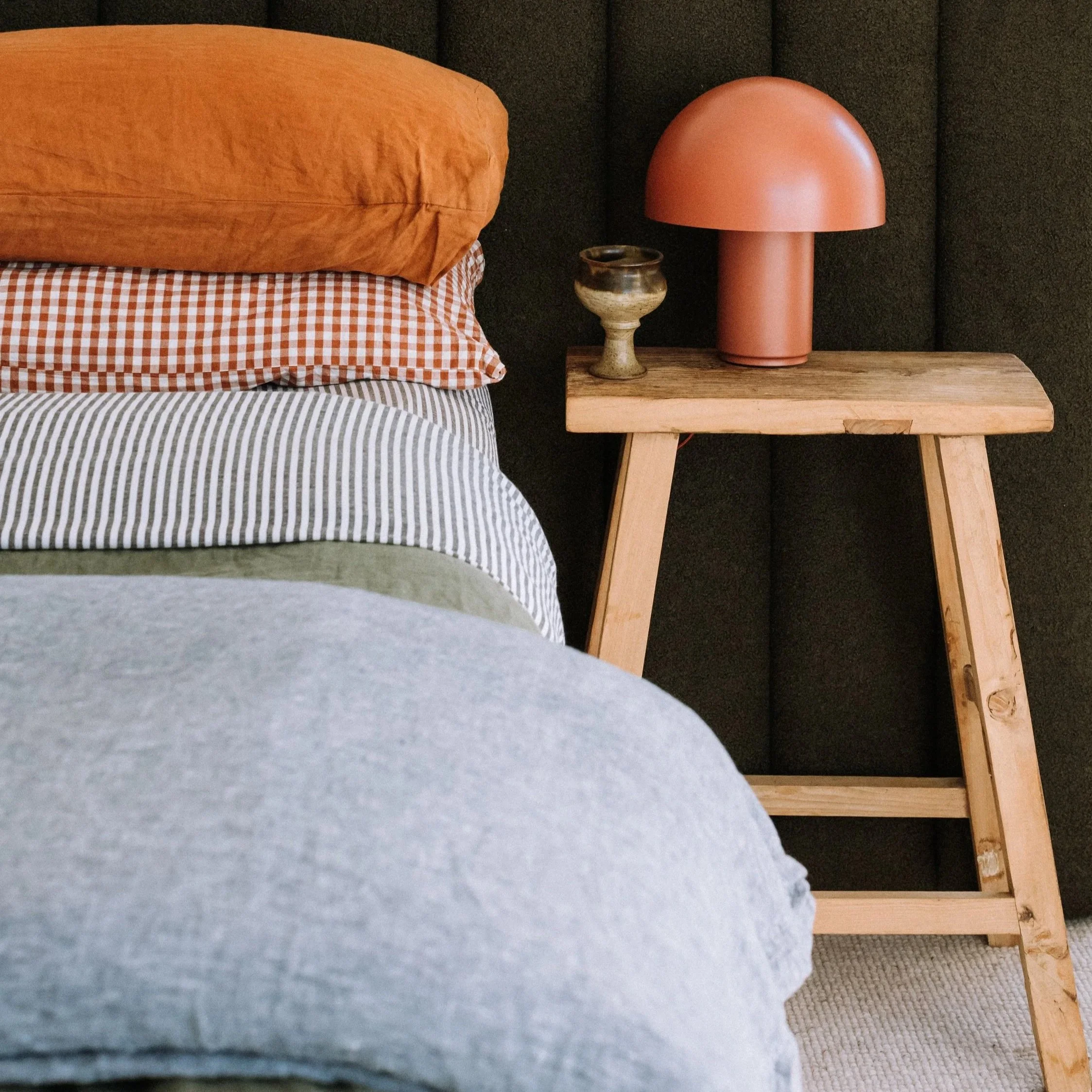 A cozy bedroom corner with a bed having multicolored striped, checkered, and solid colored pillows, and a wooden side table holding a pink mushroom-shaped lamp and a small decorative pot.