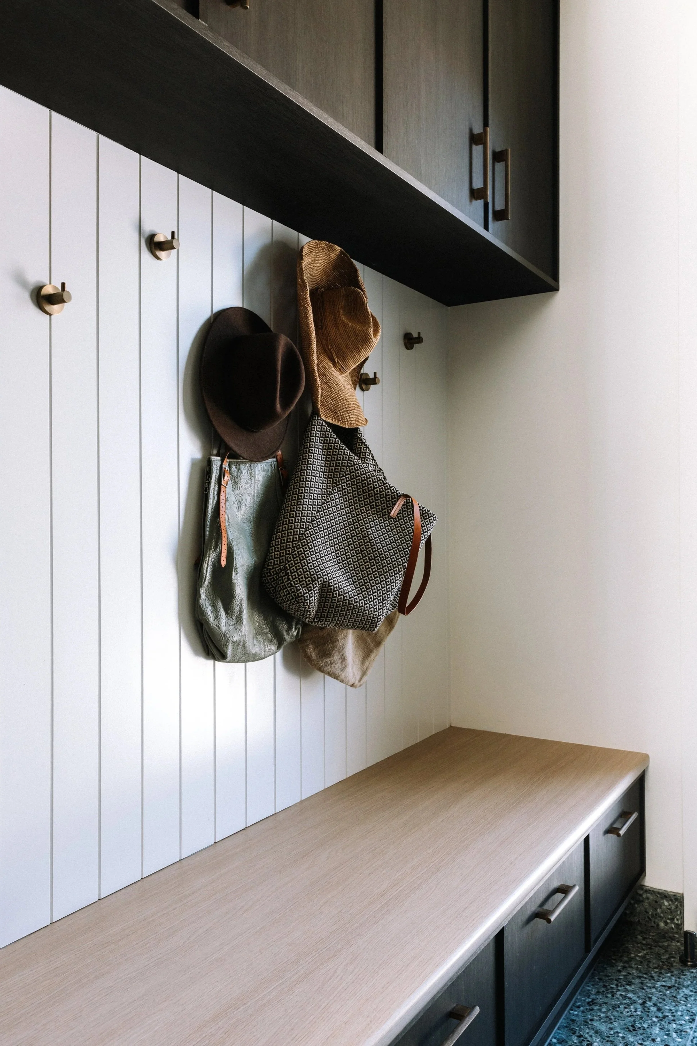 Entryway storage with bags and hats hanging on hooks, dark cabinets above, and light wood seating below.