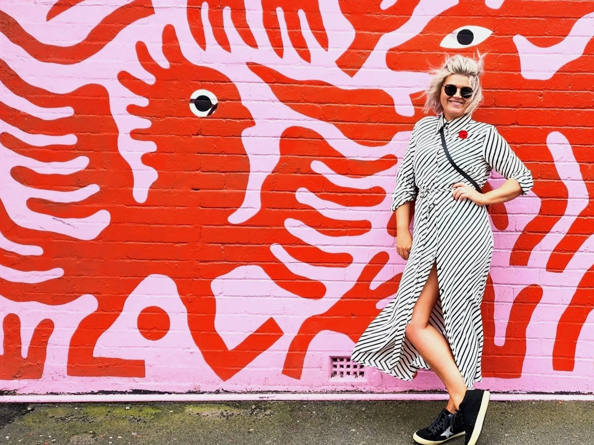 Woman with blonde hair wearing sunglasses, a black and white striped dress, and black sneakers, standing against a pink and red graffiti mural of a stylized animal face with eyes on a brick wall.