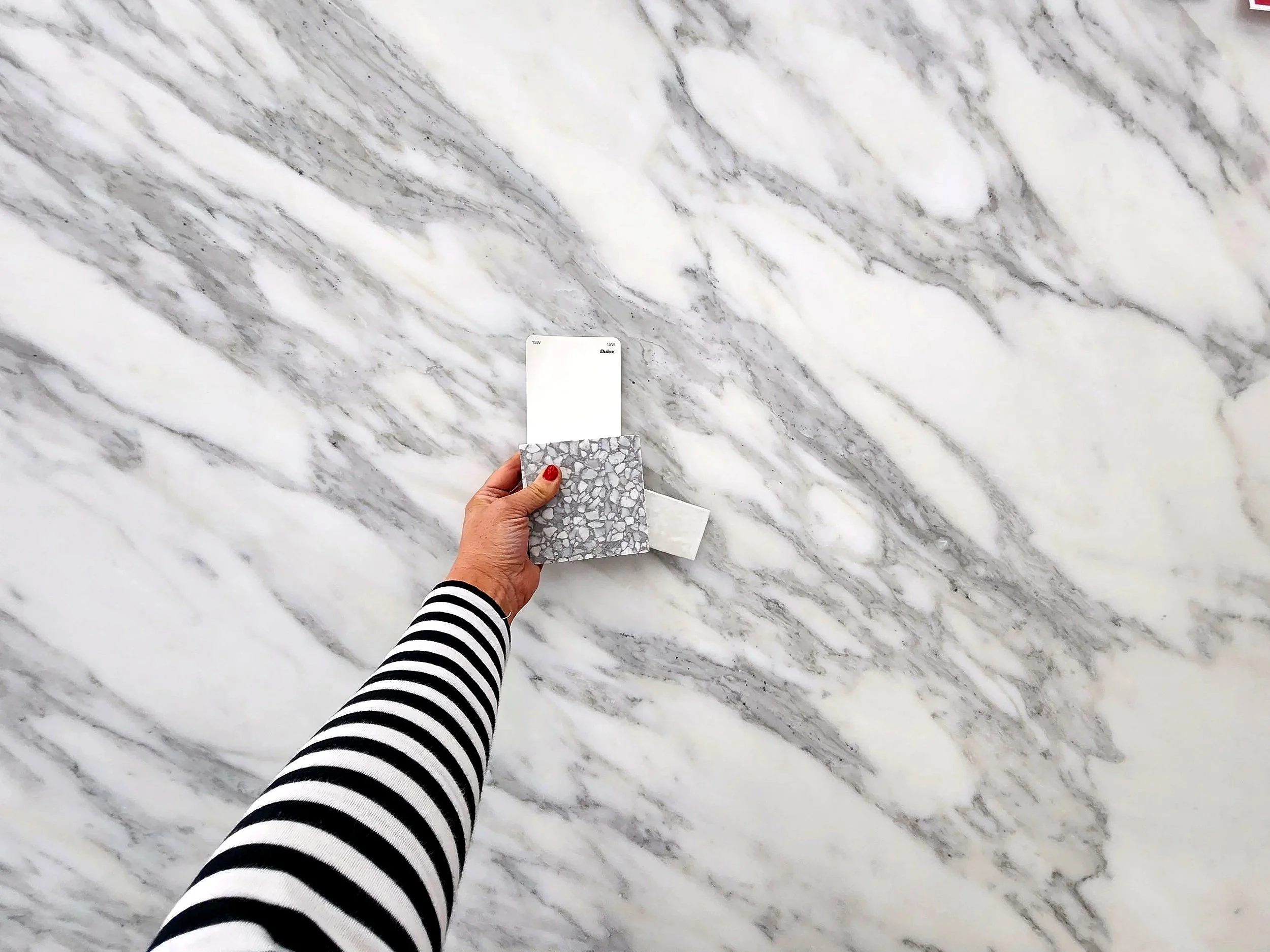 A person with red painted nails holding a sample of marble tile and a color swatch in front of a large marble wall.