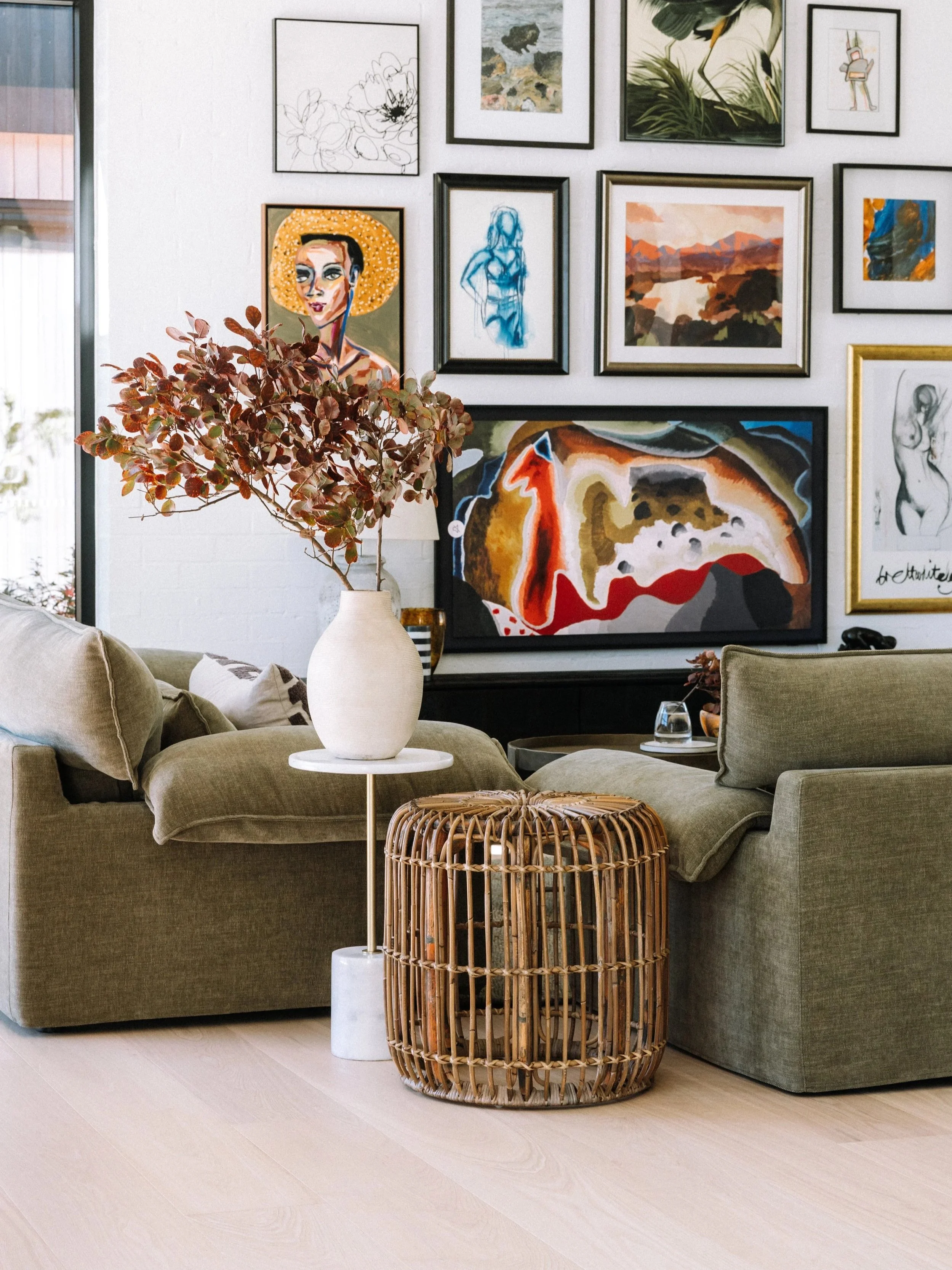 Living room with green sofa, white vase with reddish leaves, rattan side table, and gallery wall of framed abstract and figurative artwork.