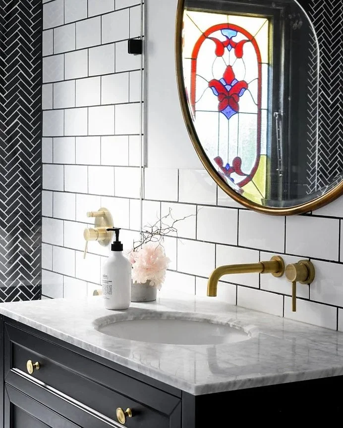 Bathroom with white subway tile walls, black lower cabinet with gold knobs, marble countertop with a built-in sink, gold faucet, and a round stained glass window with red, blue, and yellow designs.