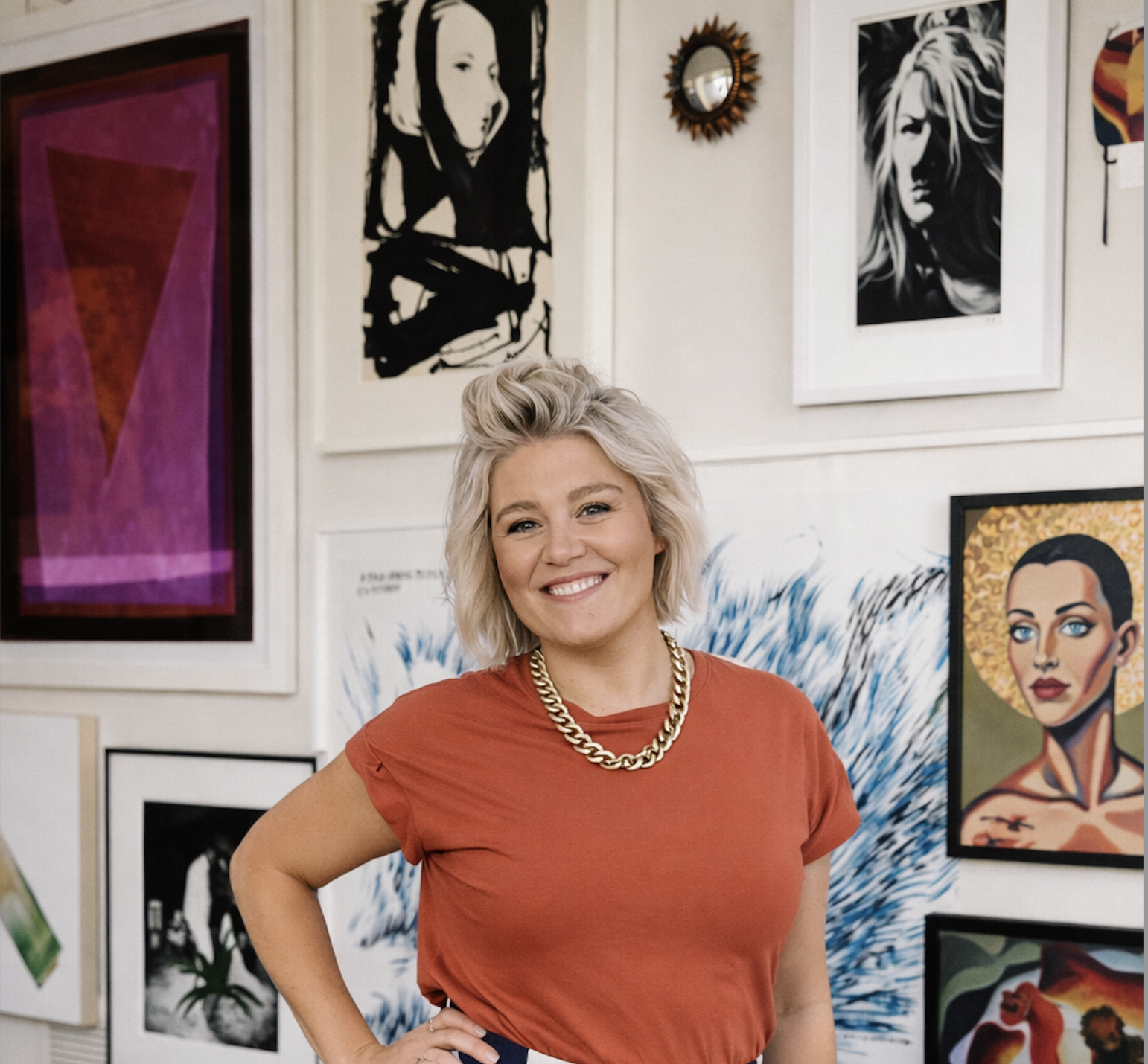 A woman with blonde hair, wearing a red top and a gold chain necklace, smiling with her hand on her hip, standing in art gallery surrounded by colorful paintings and black-and-white portraits.