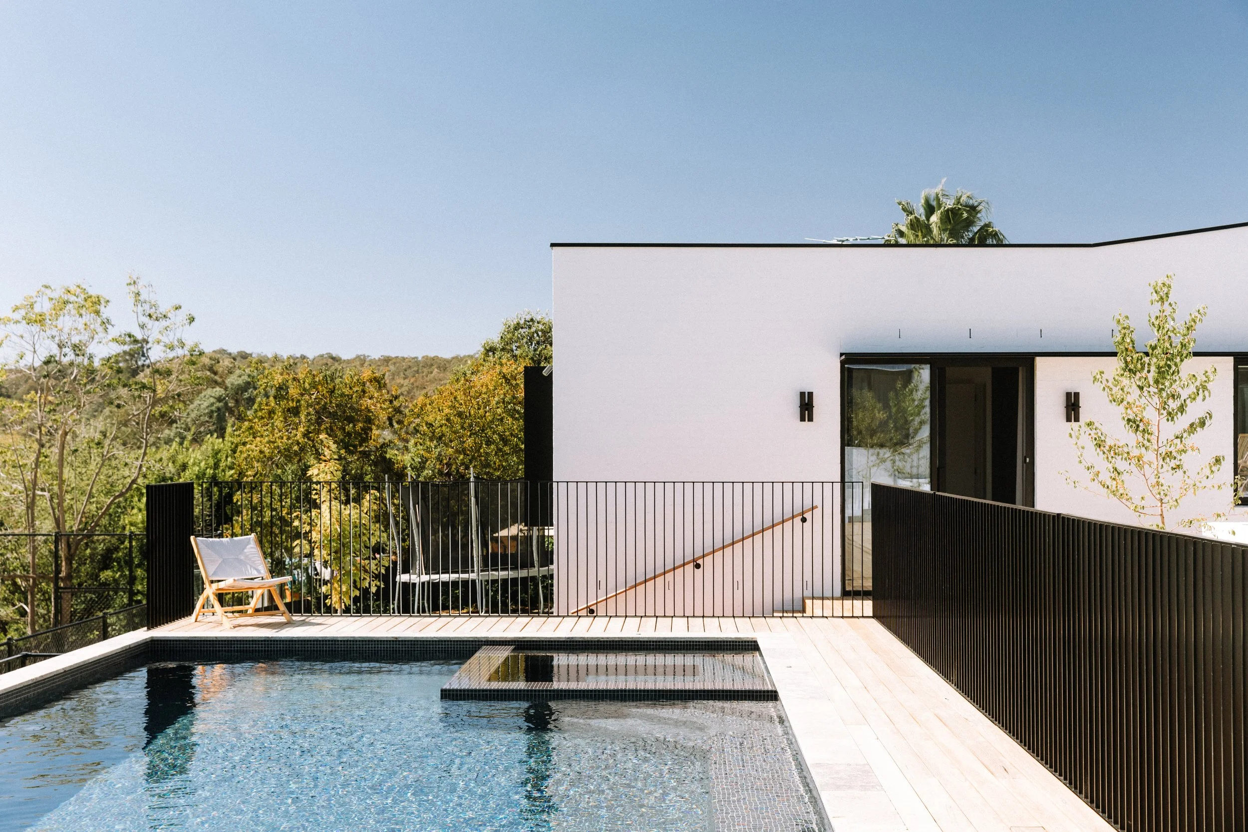 Modern house with a rooftop pool, black metal fencing, and surrounding trees under a clear blue sky.