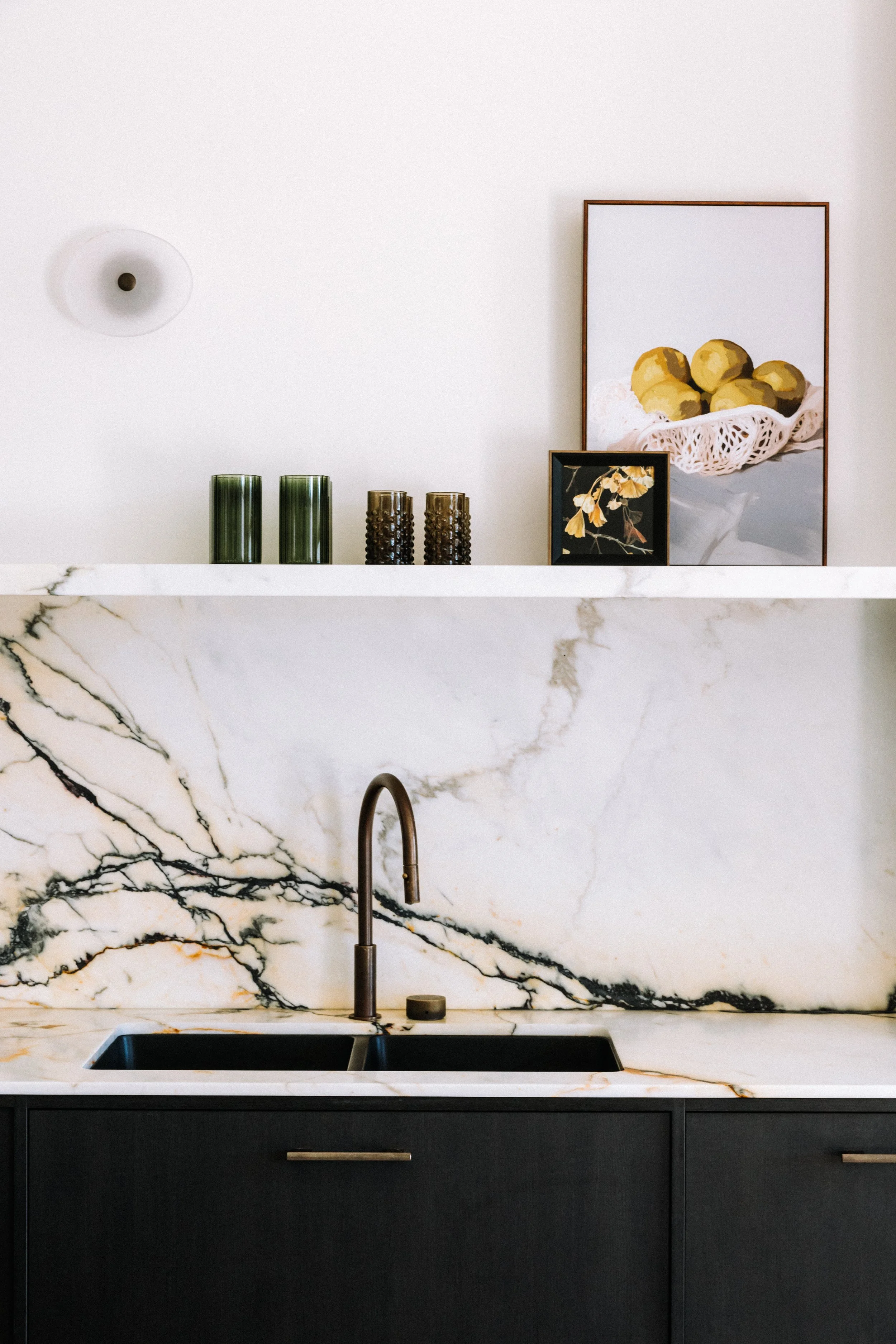 A modern kitchen with white marble countertops and backsplash, black lower cabinets, a bronze faucet, decorative vases and framed art on the wall.