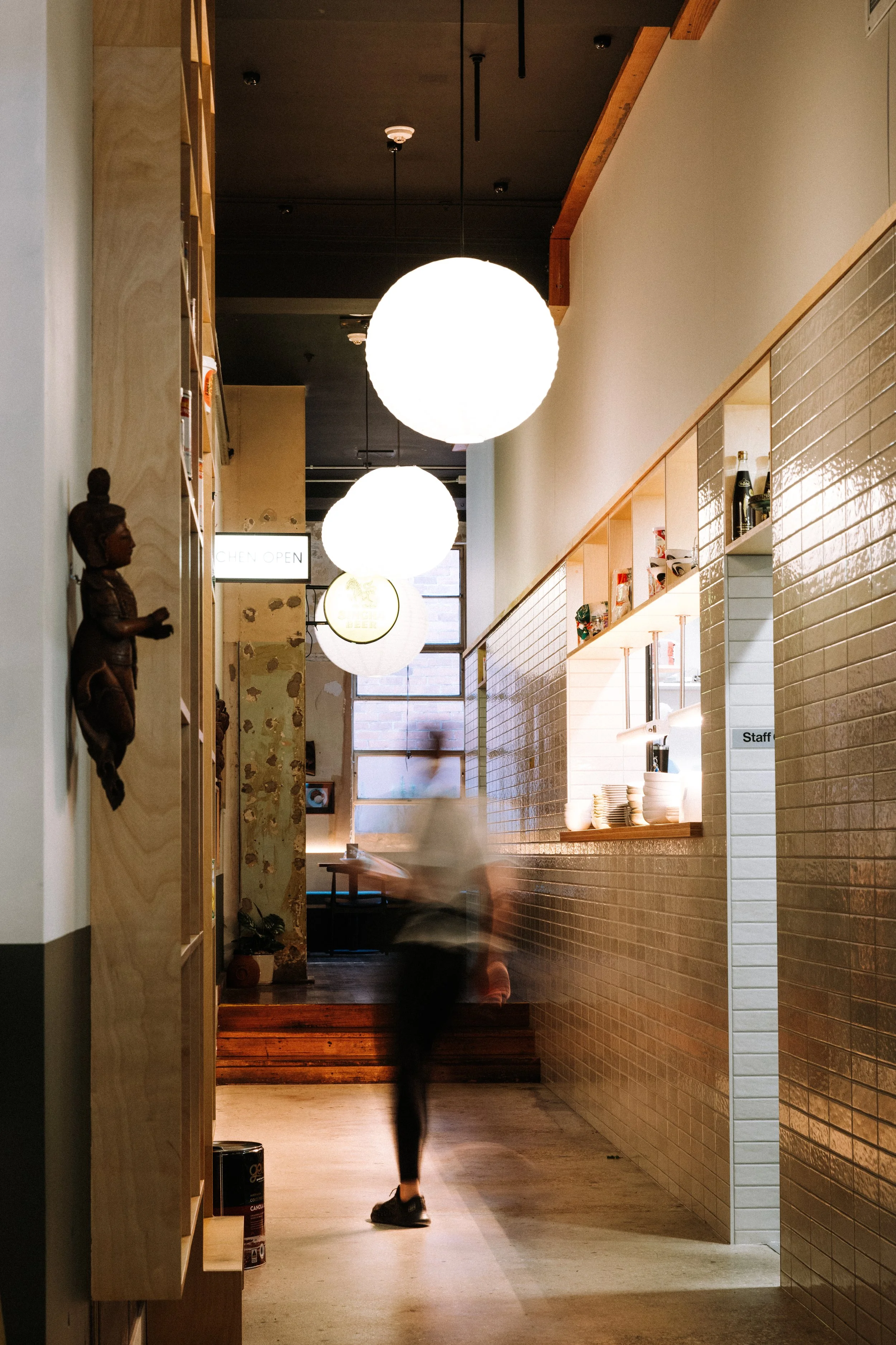 A blurred person walking in a modern restaurant or cafe interior with hanging round lights, wooden accents, and white tiled walls.