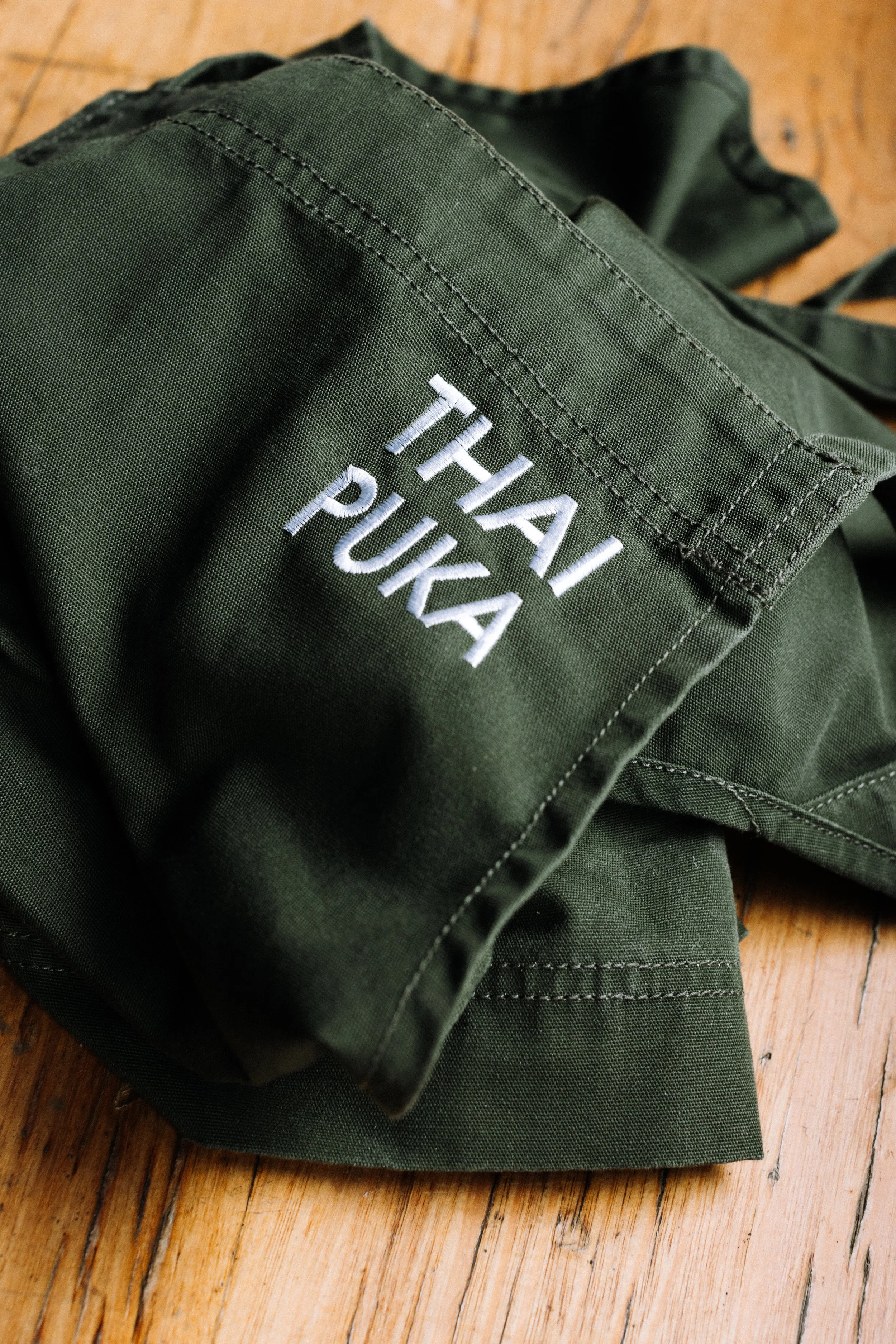 A green falcon jacket with white embroidered text that says "THAI PUKA" on the sleeve, lying on a wooden floor.