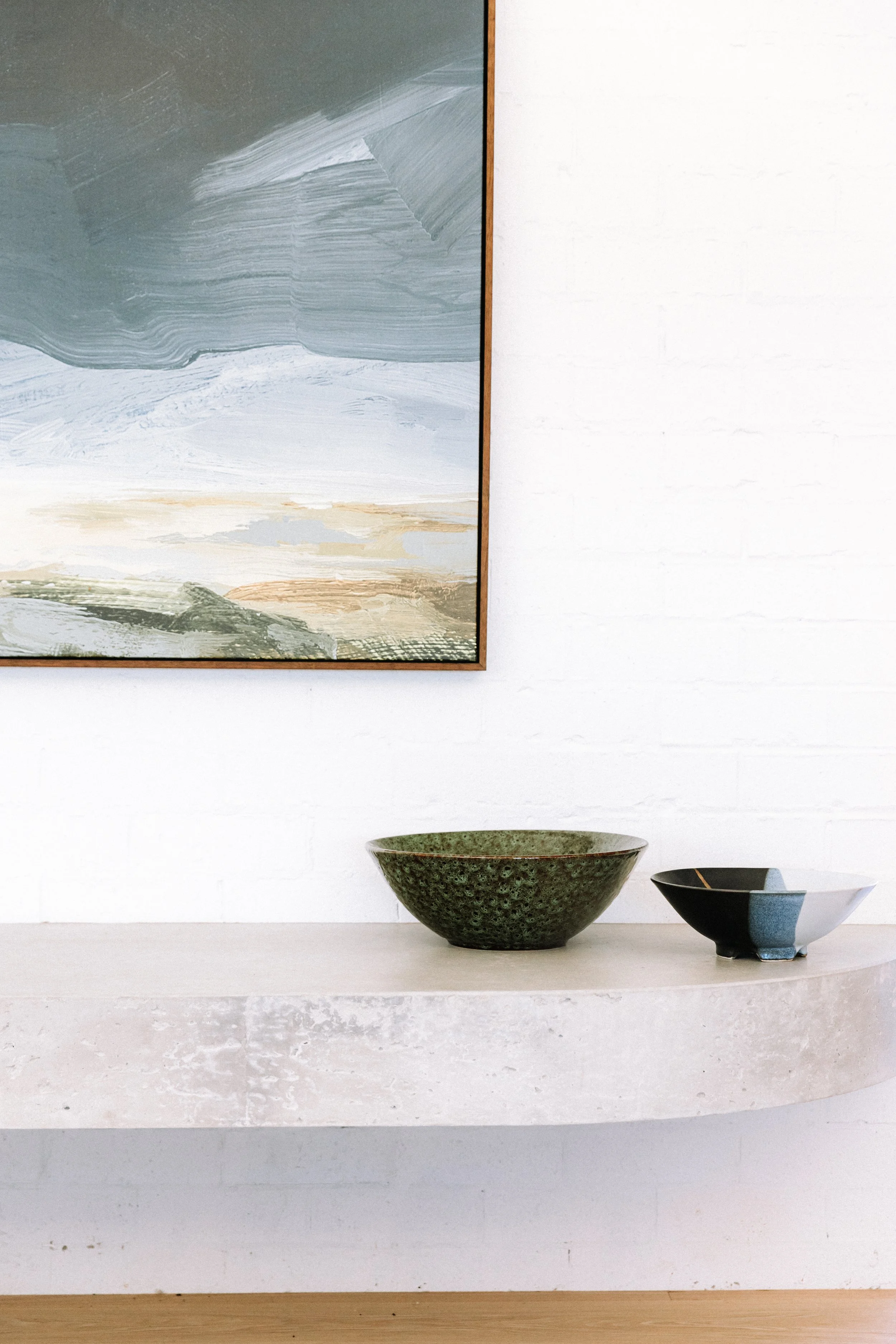 A close-up of a white concrete shelf holding two decorative bowls, one green and textured, and the other black and white with a gold line, in front of a white brick wall with a large abstract landscape painting.