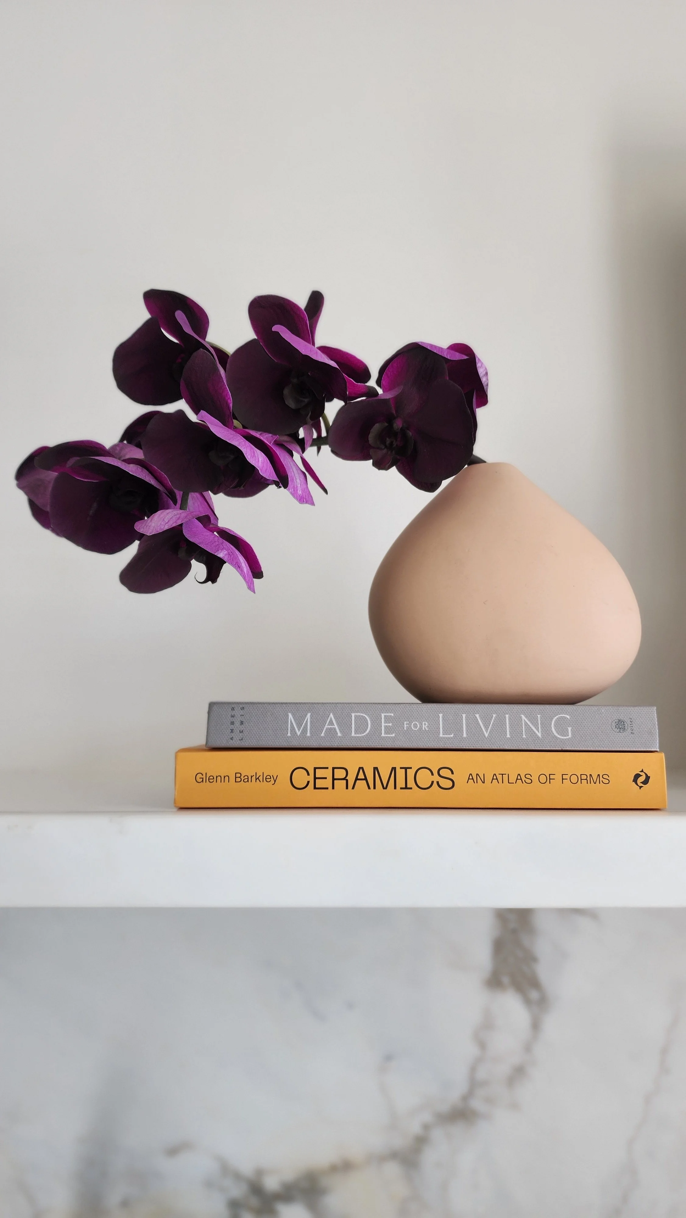 A stack of two books, one gray titled 'Made for Living' and one orange titled 'Ceramics: An Atlas of Forms,' a beige ceramic vase, and a purple orchid flower on a white shelf against a plain white background.