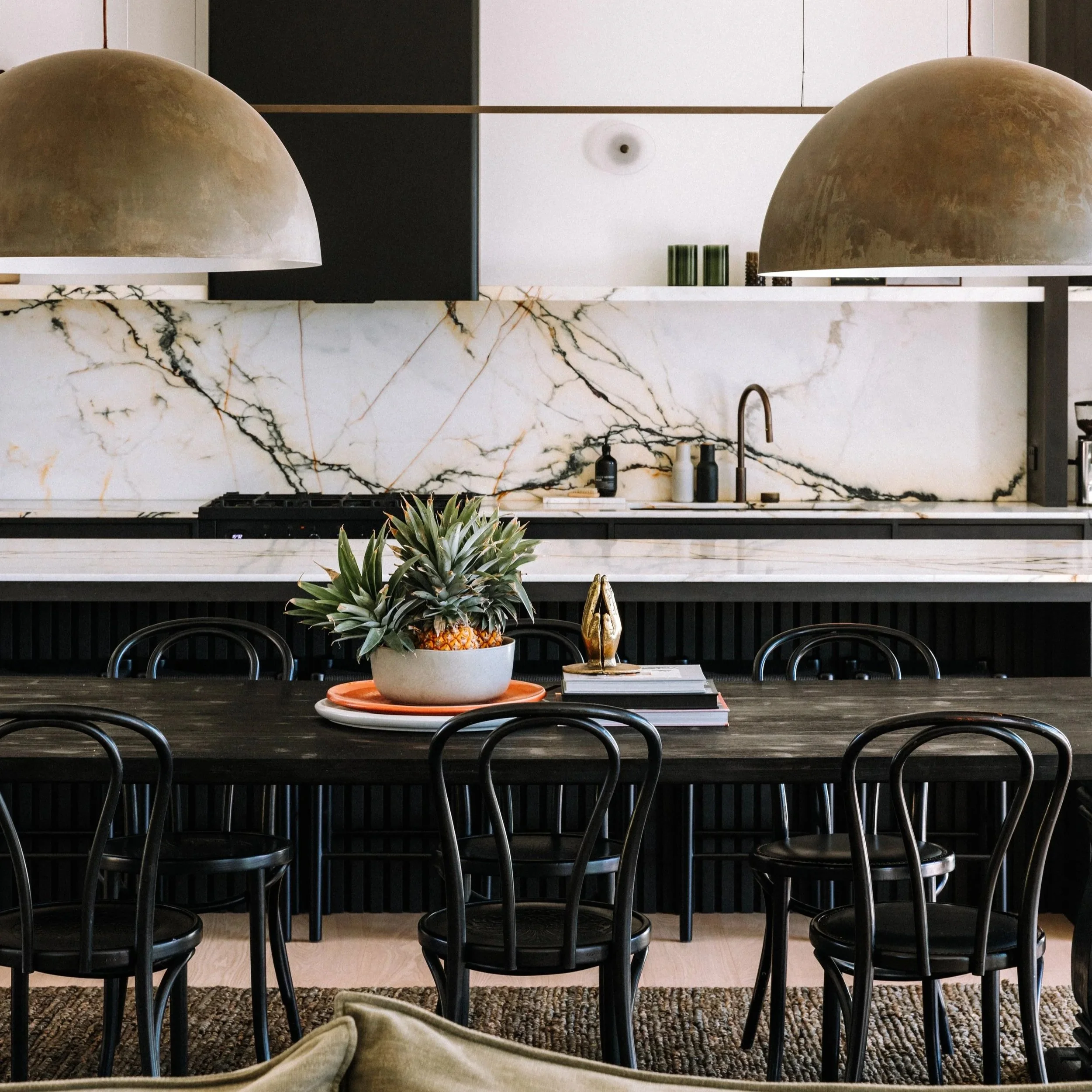 Modern kitchen and dining area with black chairs around a dark wooden table, centerpiece with pineapple plant, and gold decorative object, marble backsplash, and brown dome pendant lights.