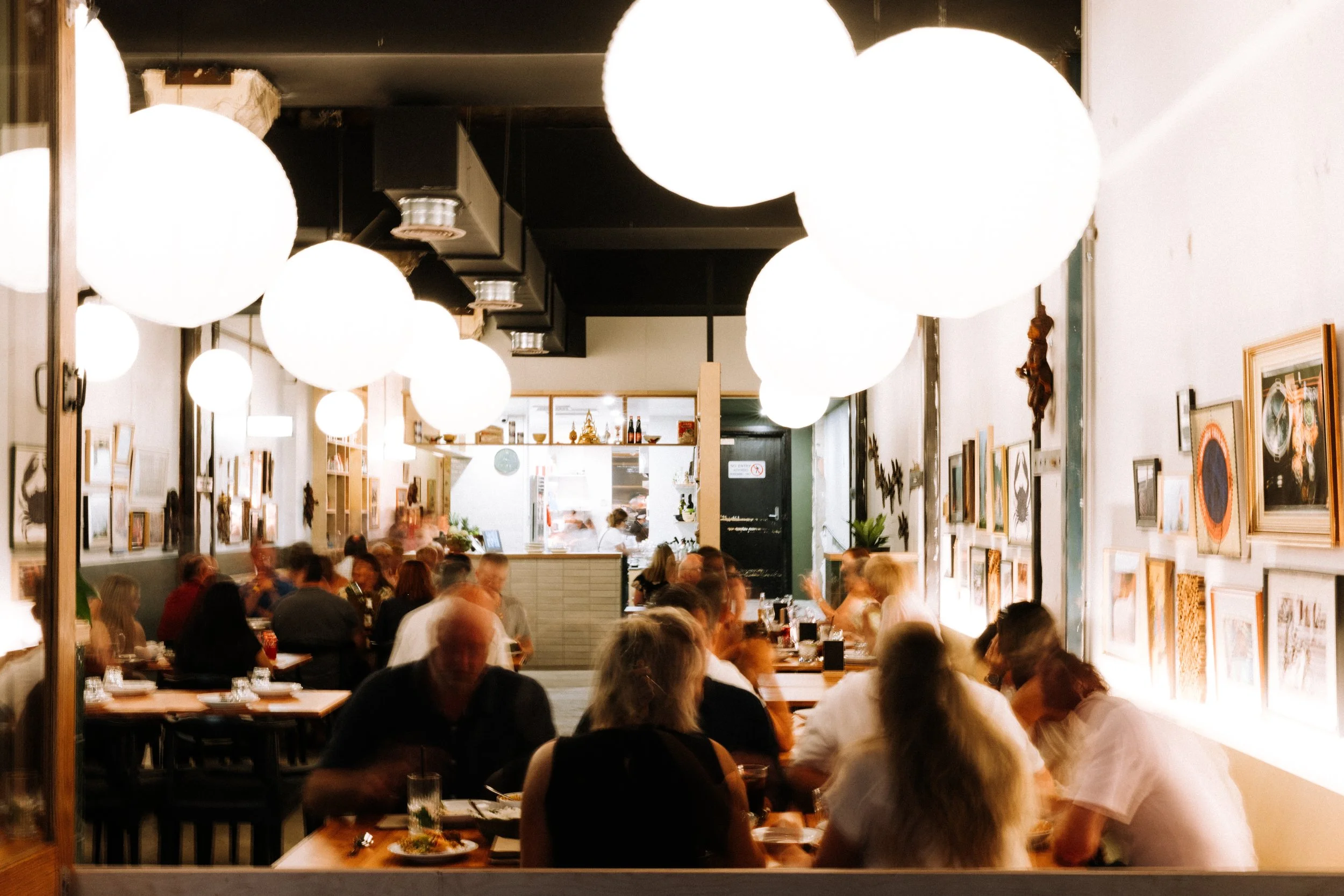 A bustling restaurant interior with hanging white spherical lights, framed artwork on the walls, and patrons seated at wooden tables enjoying their meals.