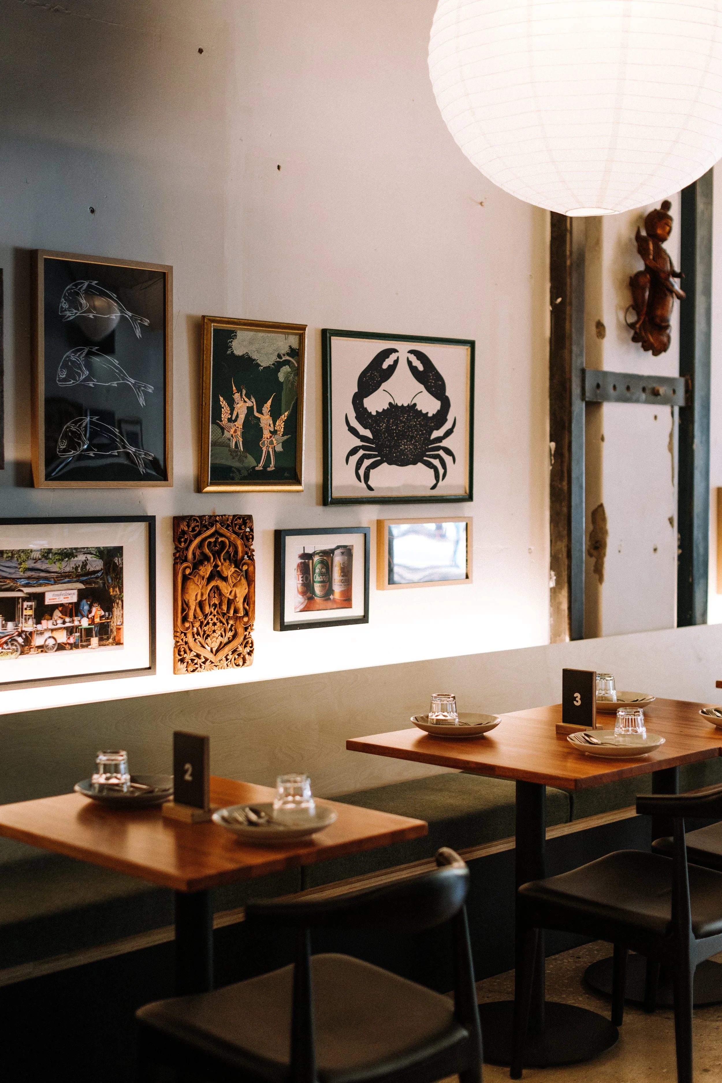 Interior of a restaurant with wooden tables set with plates and glasses, and a wall decorated with framed pictures and artwork, including a crab illustration. A round paper lantern hangs from the ceiling.