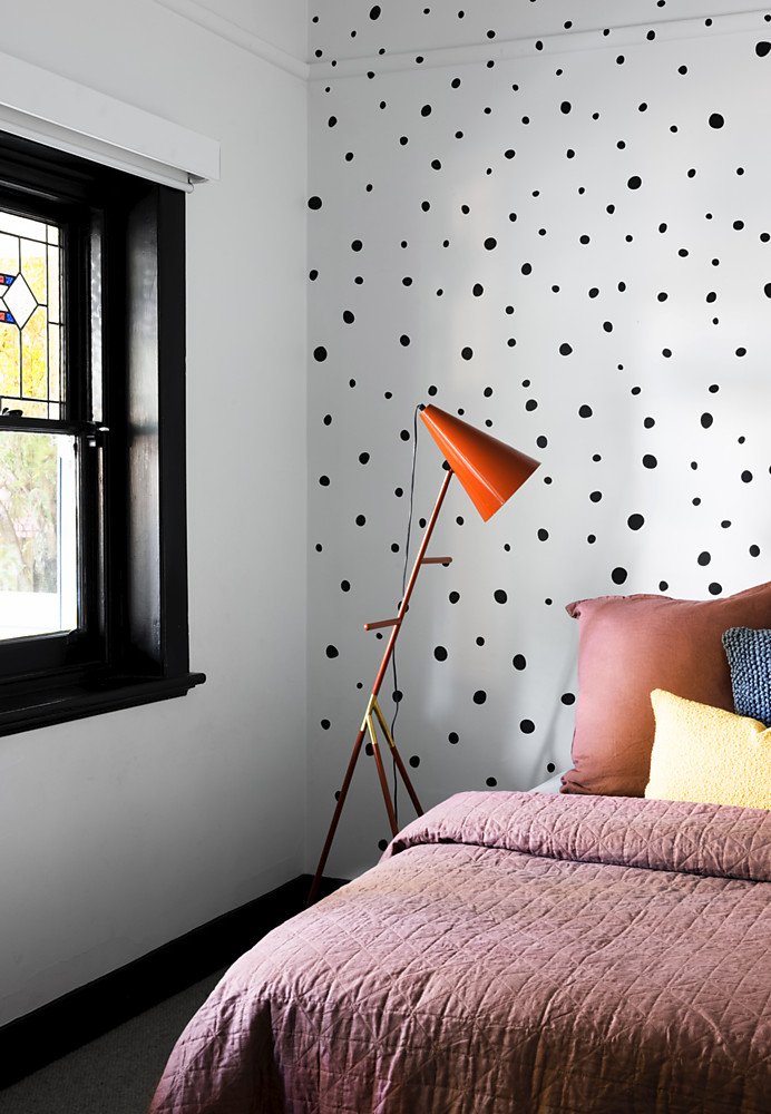 A bedroom corner with a bed featuring pink bedding, a brown pillow, and a yellow pillow, a black-framed window with stained glass, and a tall orange floor lamp against a white wall with black polka dot pattern.