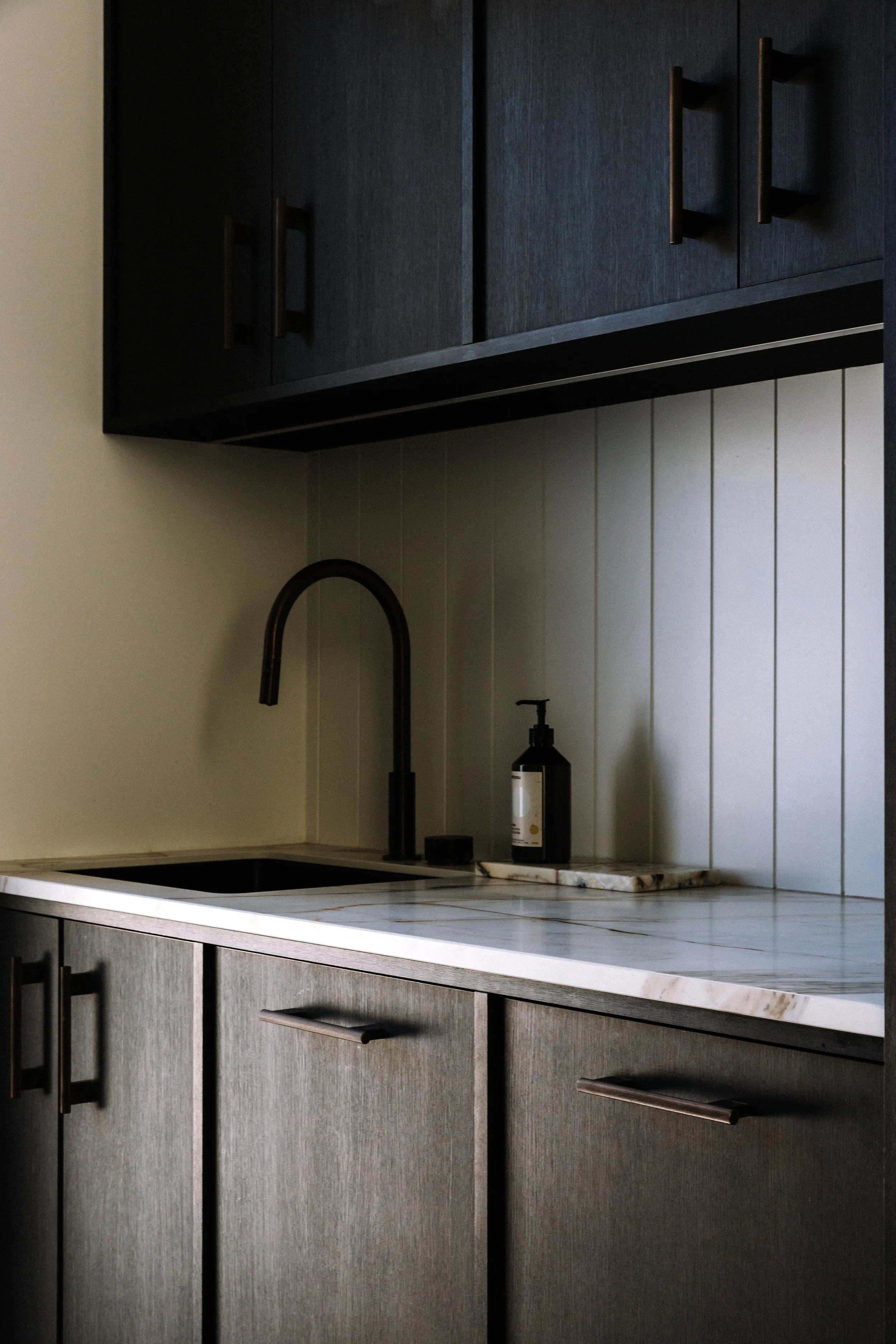 Minimalist kitchen with dark upper cabinets, gray lower cabinets, a black faucet, a soap dispenser, and a white marble countertop with subtle veining.