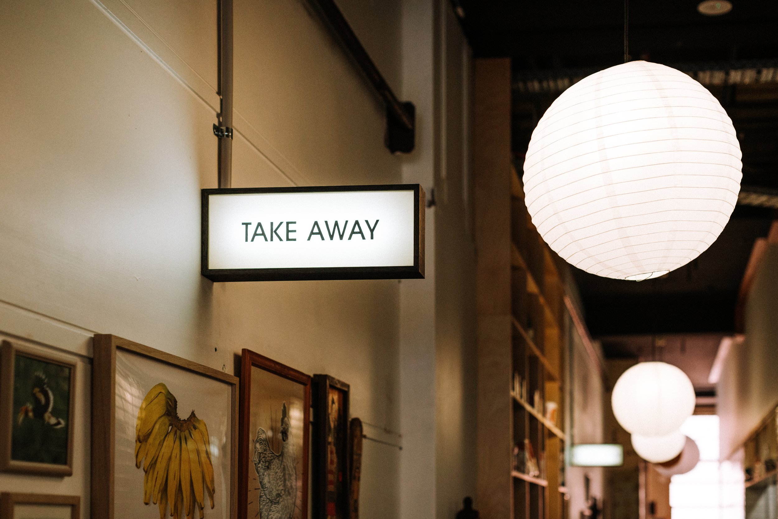 A white illuminated sign on a wall with the text "TAKE AWAY", hanging in a cozy indoor space. To the right, white paper lanterns hang from the ceiling, creating a warm ambiance. There are framed pictures on the wall below the sign.