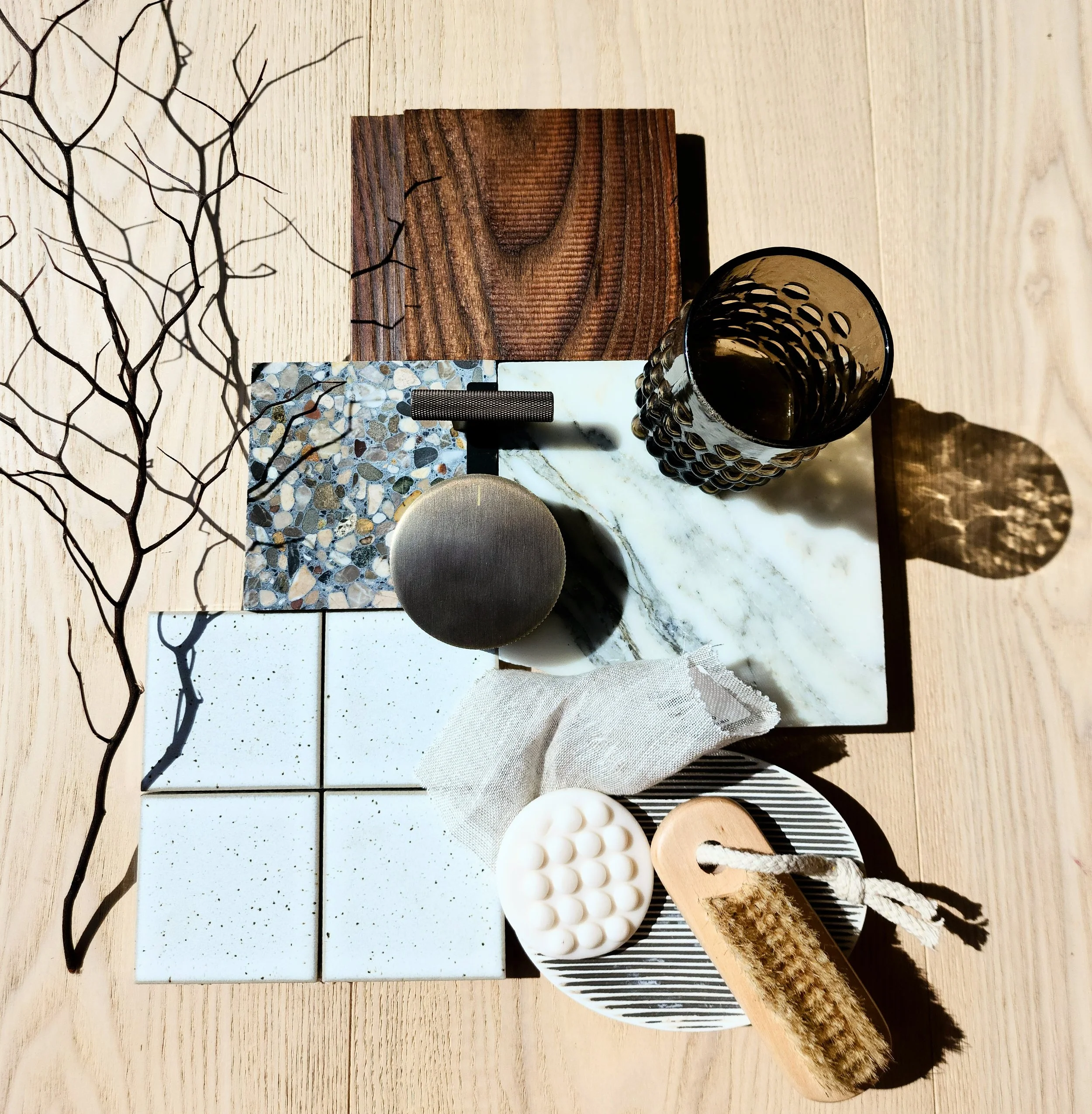 A collection of various household items arranged on a light wooden surface, including tiles, a marble slab, a glass, a soap dish, a cloth, a wooden brush, and decorative branches.