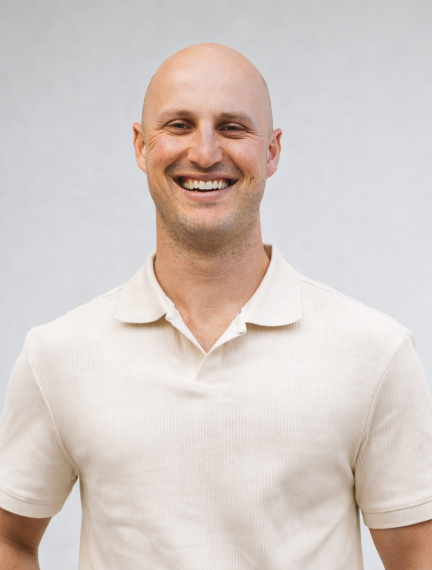 A smiling bald man wearing a light-colored polo shirt, standing against a plain background.