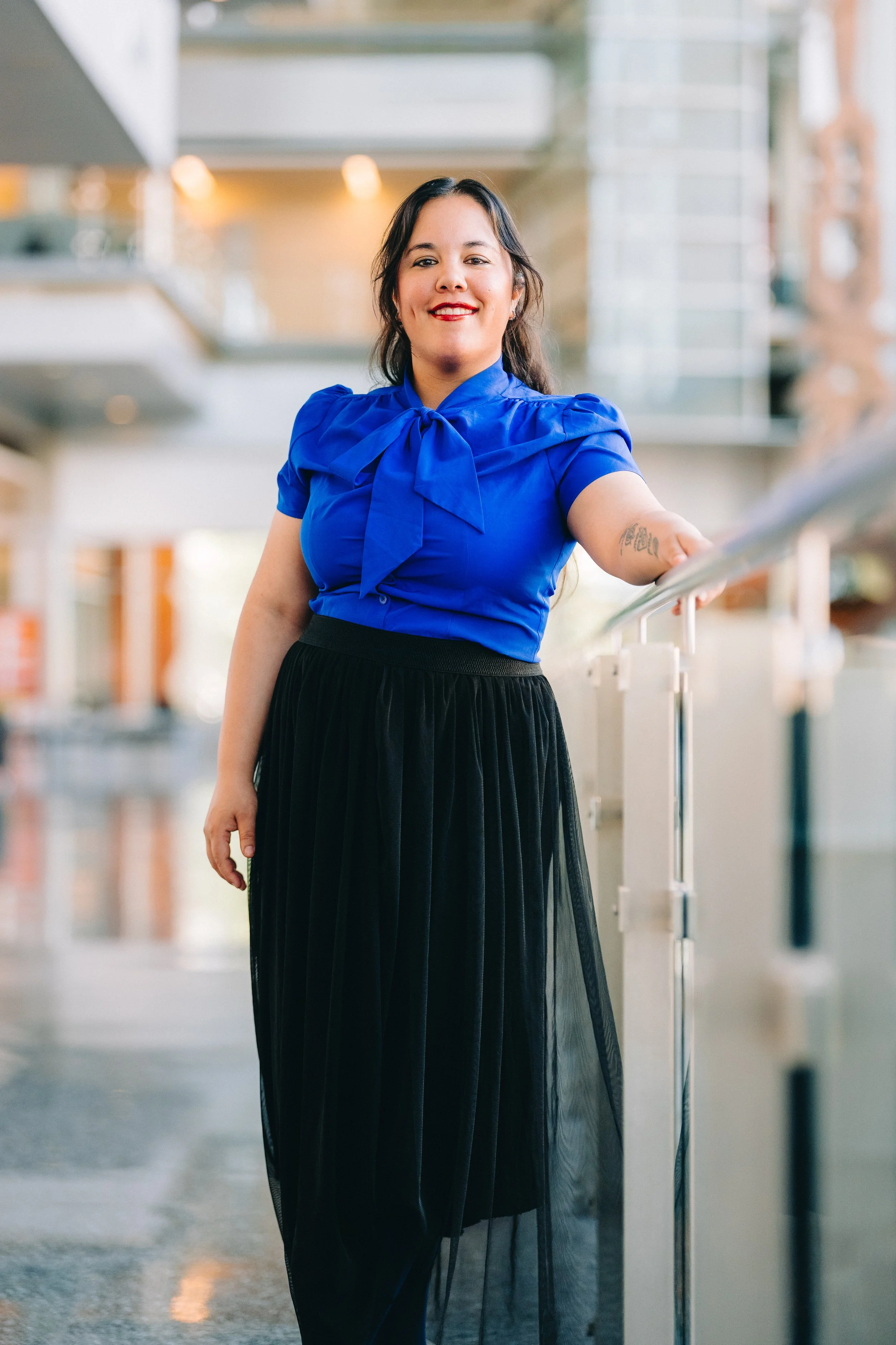A woman with dark hair, wearing a royal blue blouse with a bow tie detail and a black pleated skirt, smiling and leaning against a railing in an indoor setting with modern architecture and soft lighting.