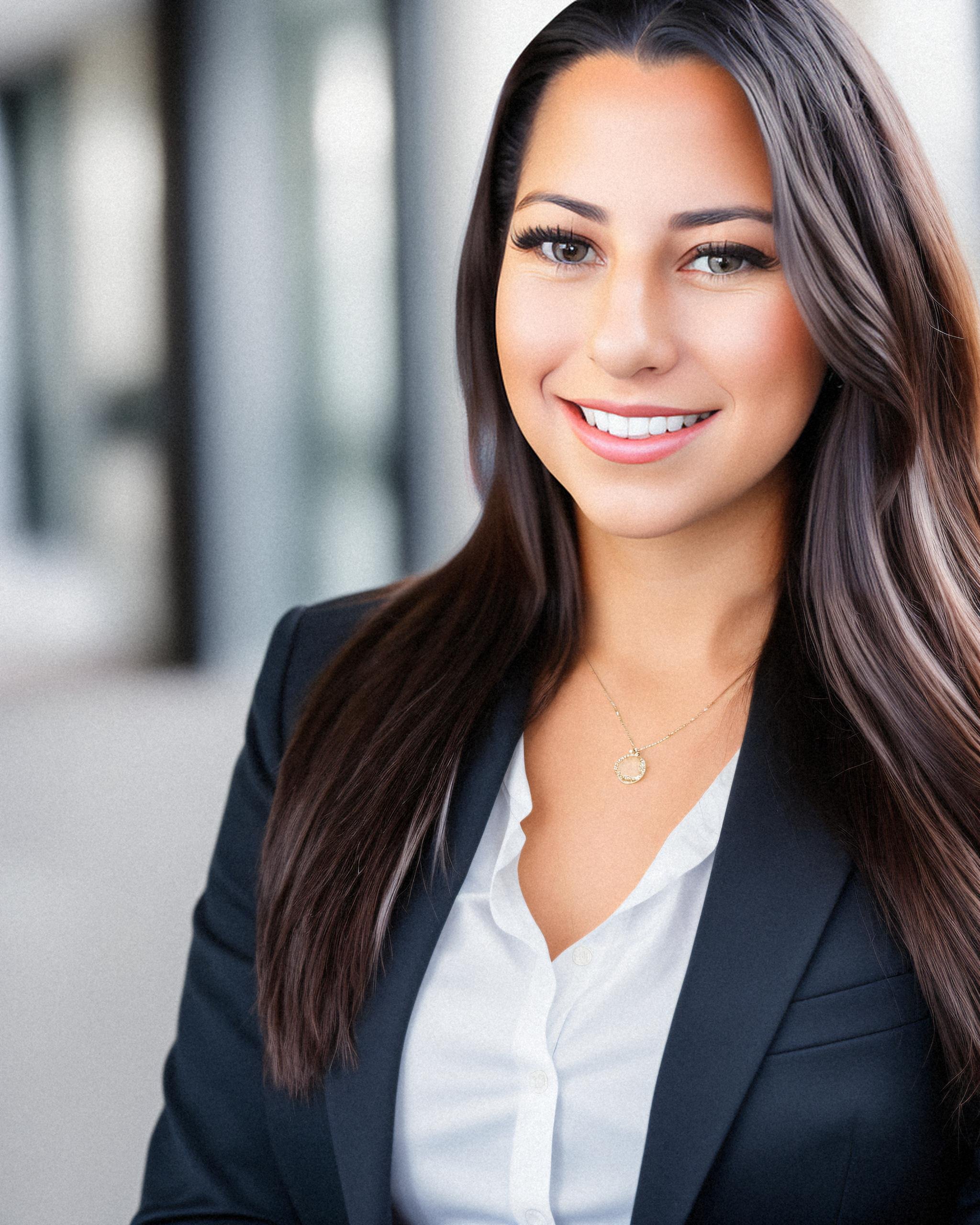 A professional woman with long dark hair wearing a black blazer, white shirt, and gold necklace, smiling confidently against a blurred office background.