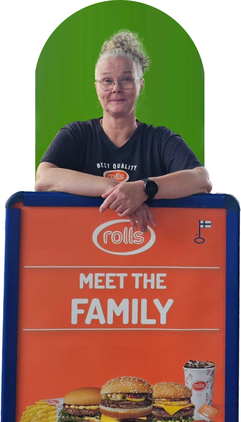 A woman with glasses and curly gray hair, wearing a black shirt and a watch, standing behind an orange sign with the Rolls logo. The sign reads 'Meet the Family' and features images of various burgers, fries, and a drink, with a green background behind her.