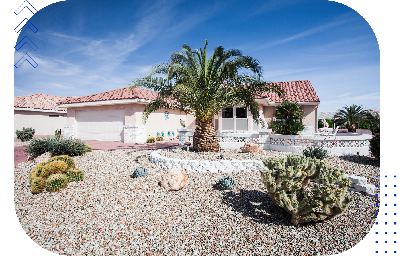 Single-story house with a red tile roof, white walls, a double garage, and desert landscaping featuring cacti, succulents, a palm tree, and rocks under a blue sky.