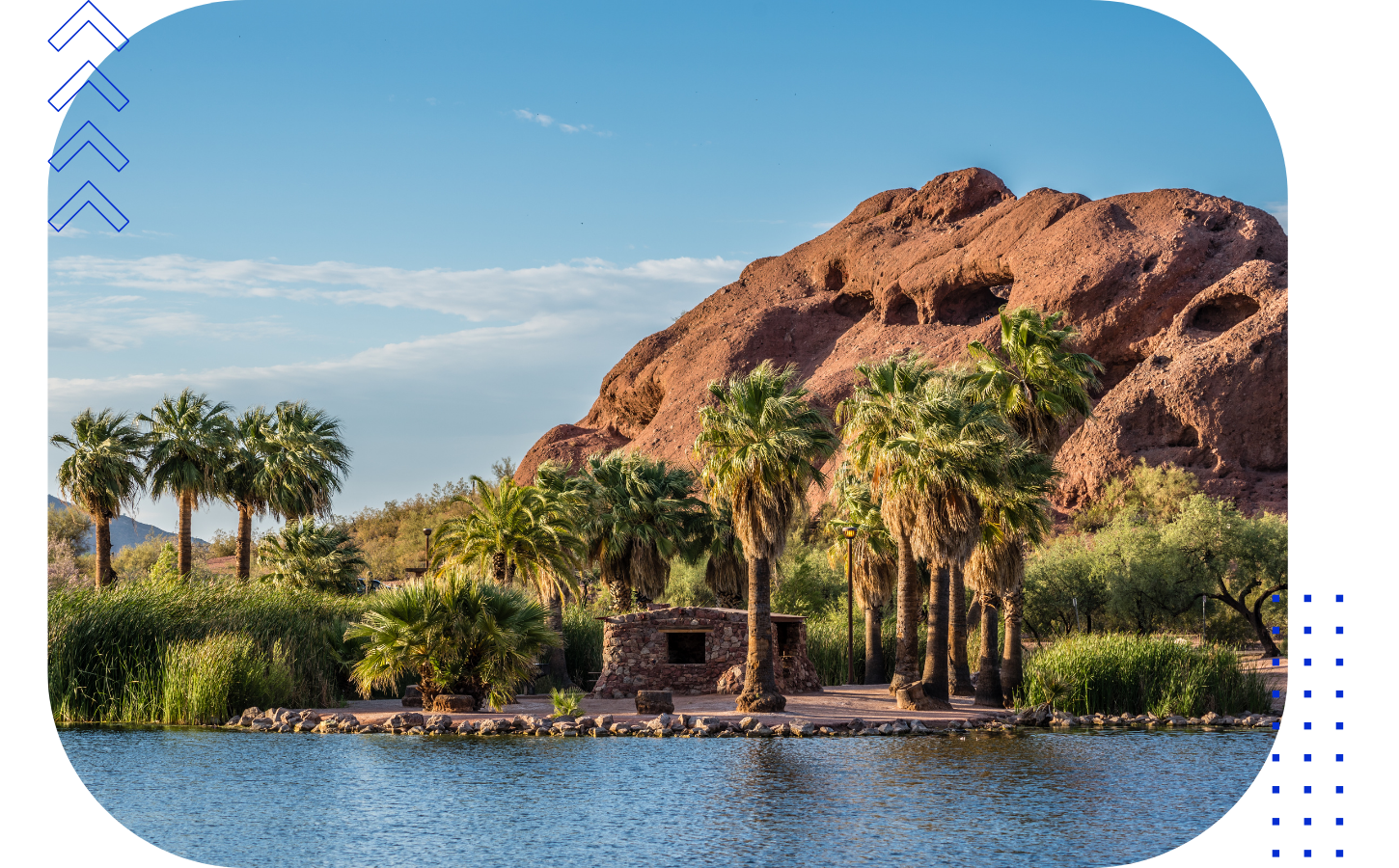 A desert landscape with a large rocky hill in the background, palm trees, a small stone building, and a water body in the foreground.