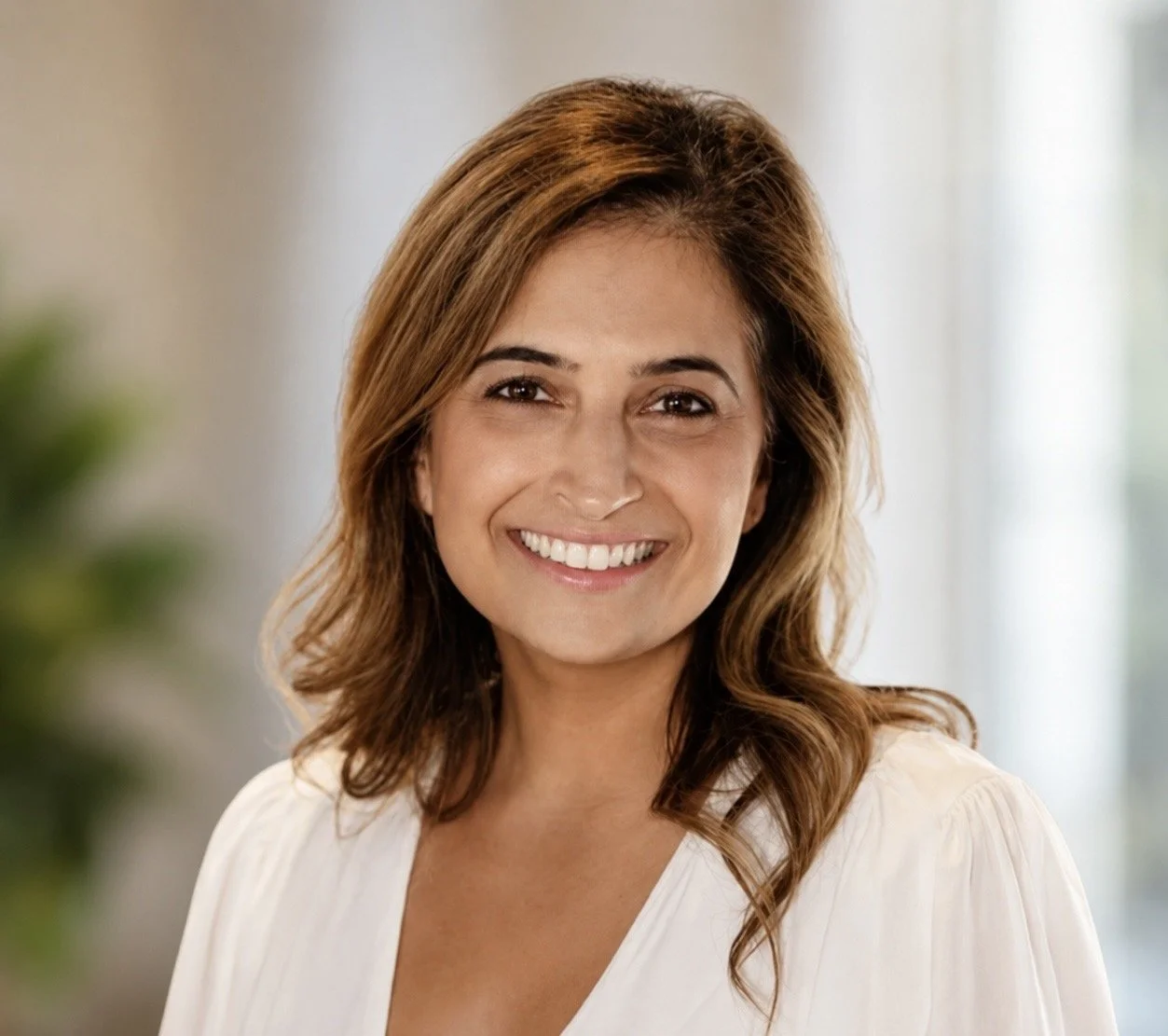 A portrait of a woman with shoulder-length wavy brown hair, smiling with white teeth, wearing a white top, and standing indoors with a blurred background.