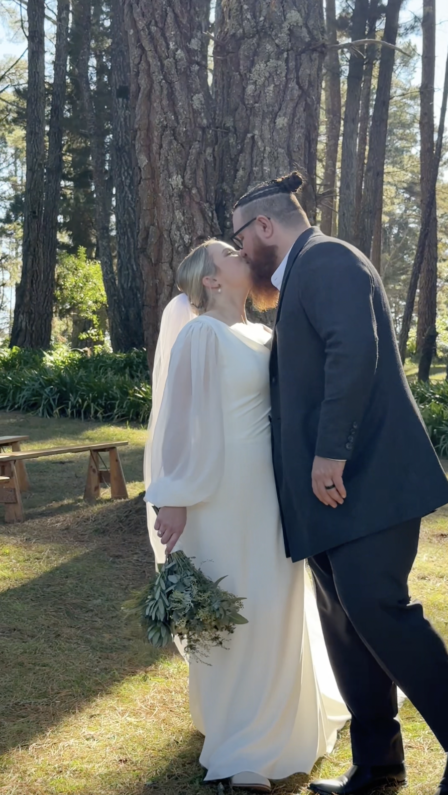 A couple, a woman in a white gown and a man in a dark suit, share a kiss outdoors in front of a large tree, during what appears to be a wedding.
