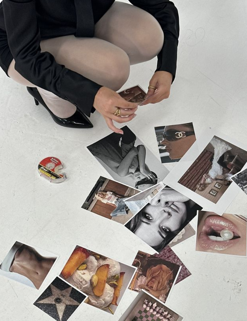 A person sitting on the floor with crossed legs, wearing a black blazer, nude stockings, and black heels, holding photographs with various images, including a black and white portrait of a woman, a woman in sunglasses, a bedroom, a face close-up, and a tongue with a pill. There are additional photographs and a roll of Scotch tape scattered on the white floor.