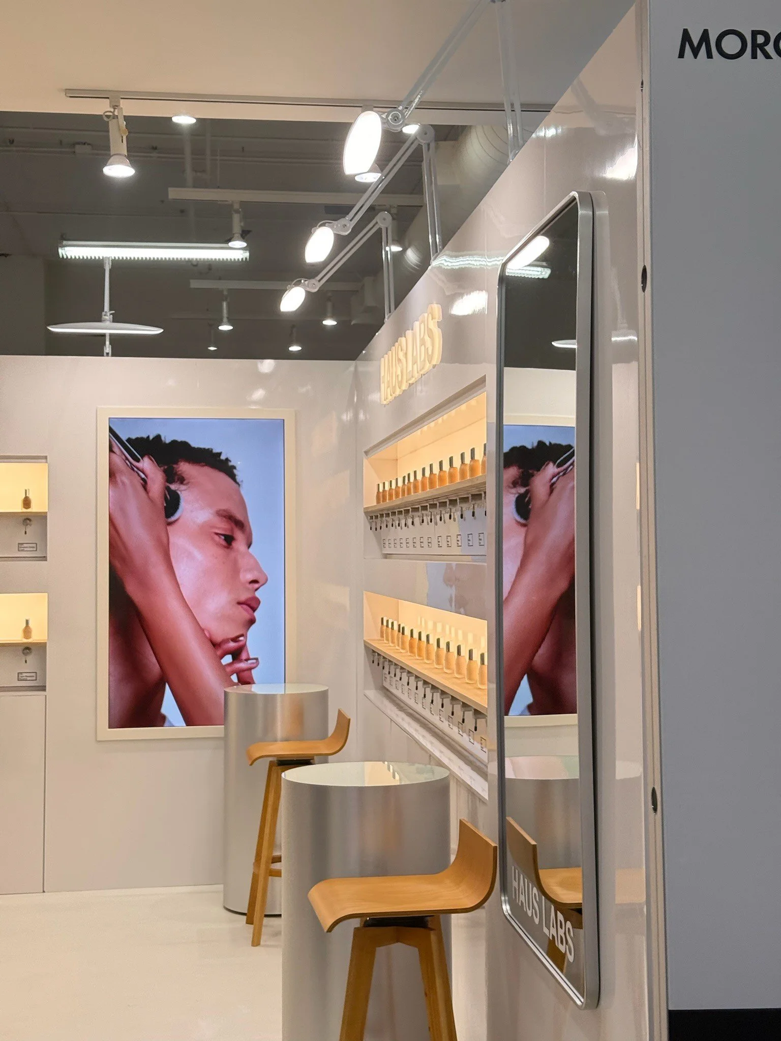 Interior of a store or showroom with a large advertisement featuring a woman applying skincare or makeup, shelves with small bottles, a small round table with chairs, and a sign with the words 'HAUS LABS'.