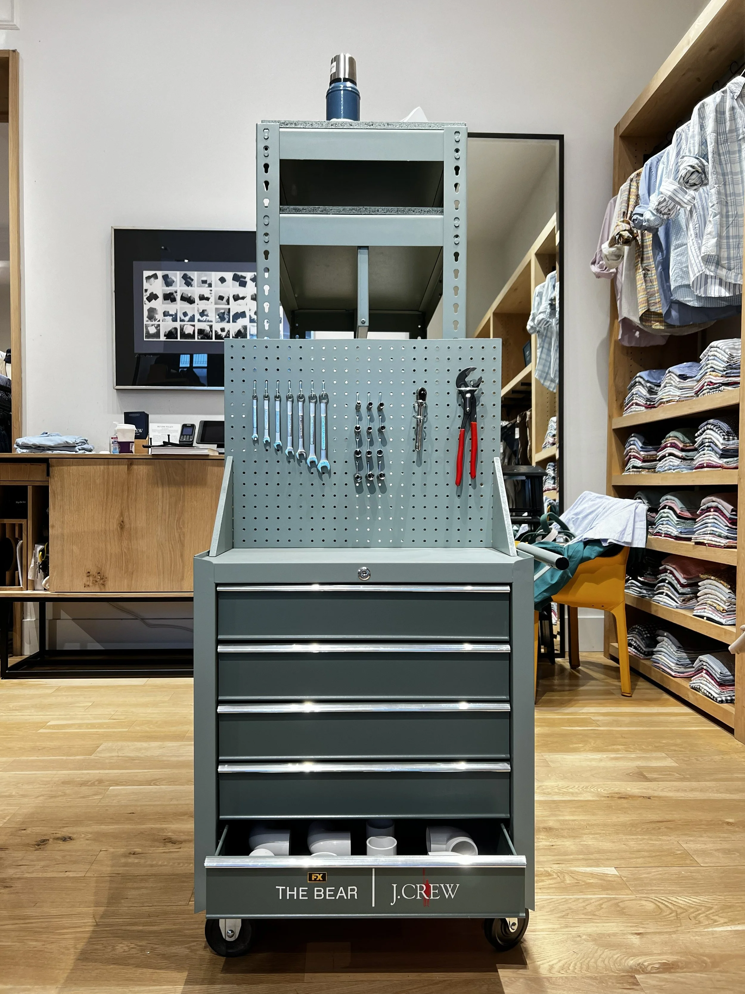 A gray tool cart with various wrenches and tools hanging on a perforated board, placed in a retail store with shelves of folded clothes on the right and a wall with a monitor and wooden countertop in the background.