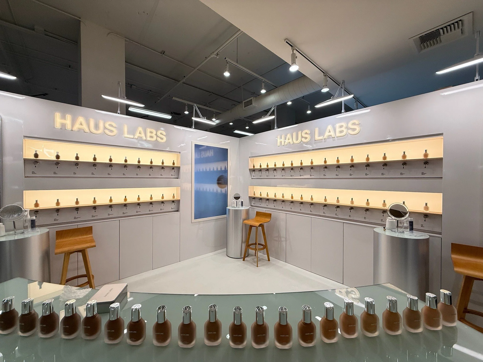 Interior of a store or showroom called 'Haus Labs,' featuring a display of small bottles arranged in rows on a clear surface, with bright lighting and minimalist modern furnishings.