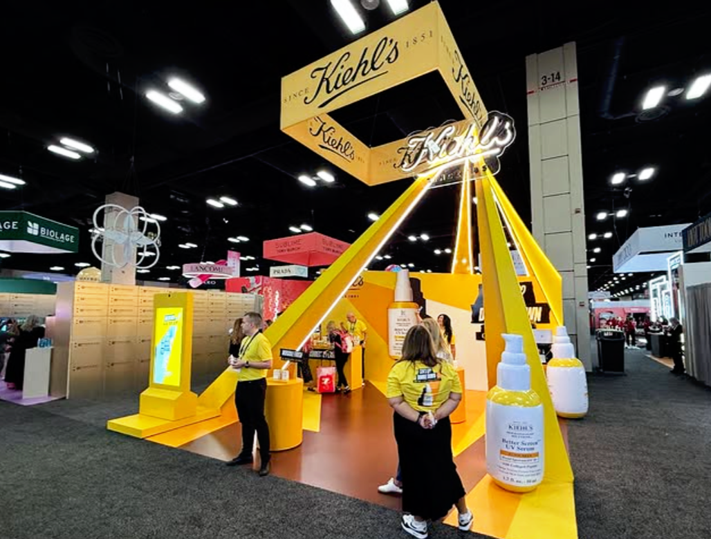 A Kiehl's promotional booth at a trade show with yellow and orange colors, large product images, and staff in yellow shirts, inside a convention center.