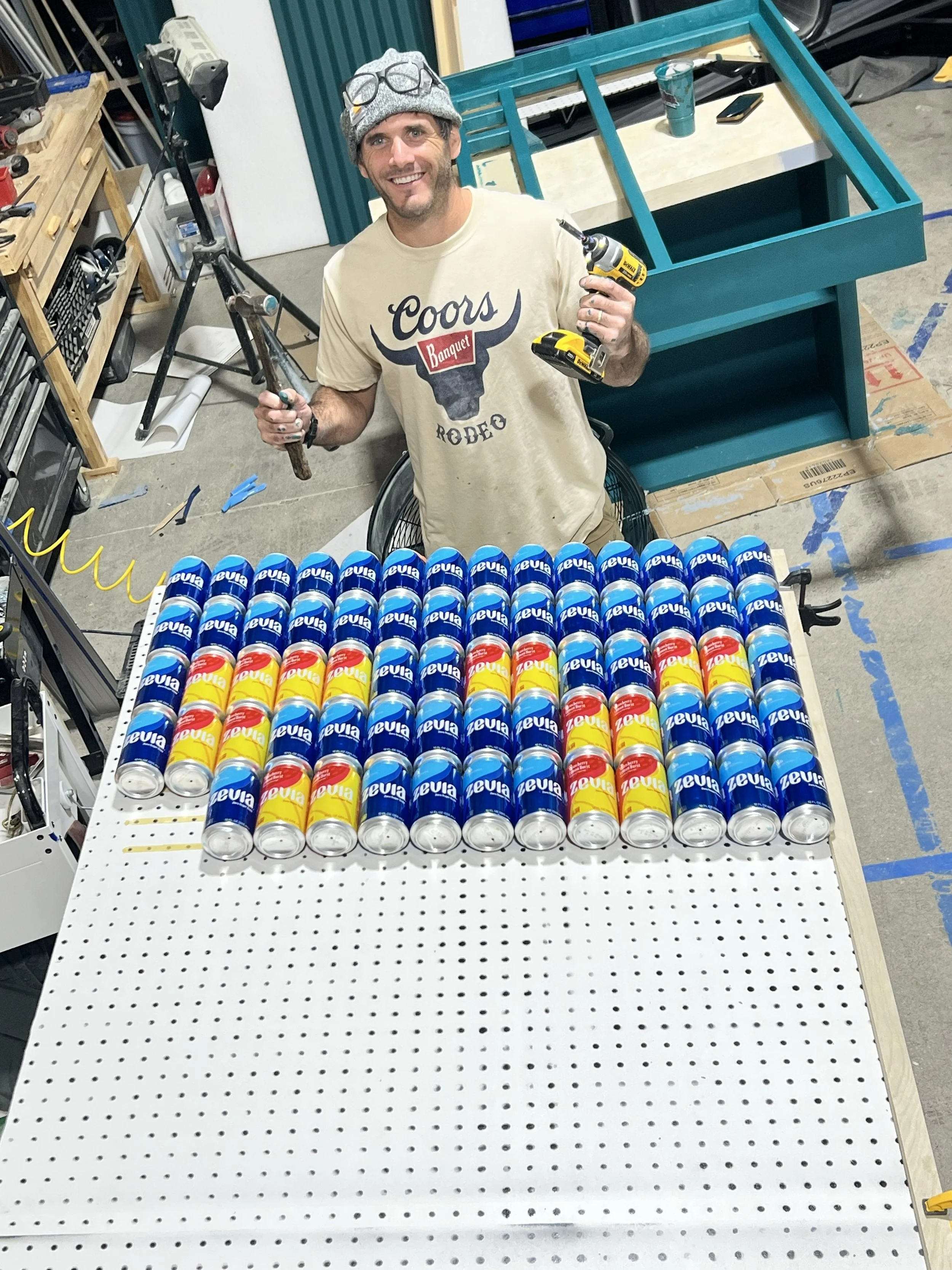 A man smiling and holding a drill and a hammer stands behind a workbench filled with cans of Zevia soda in a workshop. There is a partially assembled teal-colored furniture piece in the background.