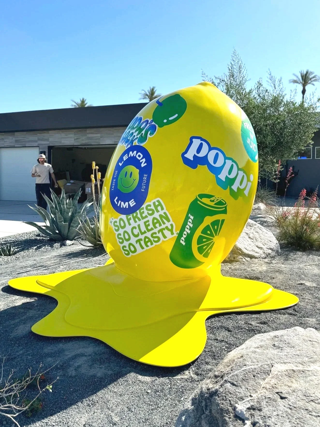 Large yellow sculpture of a lemon-flavored soda can and lemon slice with text reading 'Poppi' and 'So fresh, so clean, so tasty'. The sculpture is shaped like a lemon and is placed outdoors on a gravel surface surrounded by desert plants. A person stands in the background near a modern house with a garage.