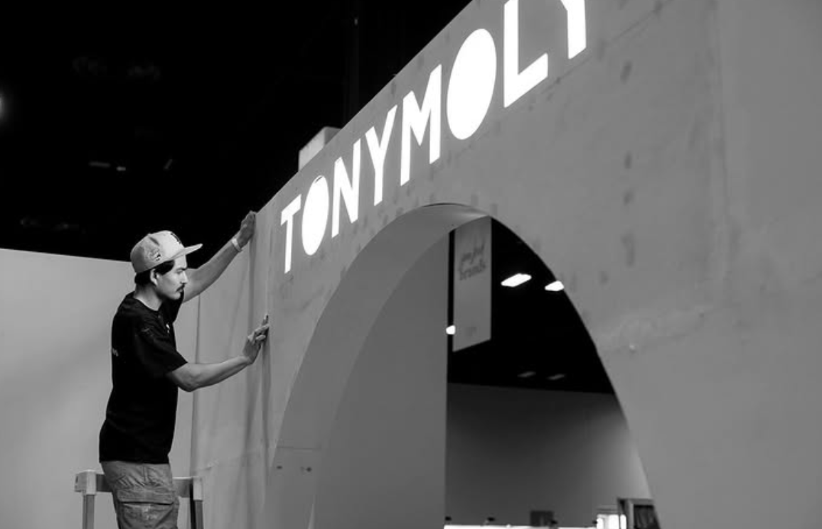 A person wearing a cap and glasses is standing on a ladder and installing or adjusting a large sign that reads 'TONY MOLY'. The sign is on a wall with an arch shape, and the scene appears to be indoors.