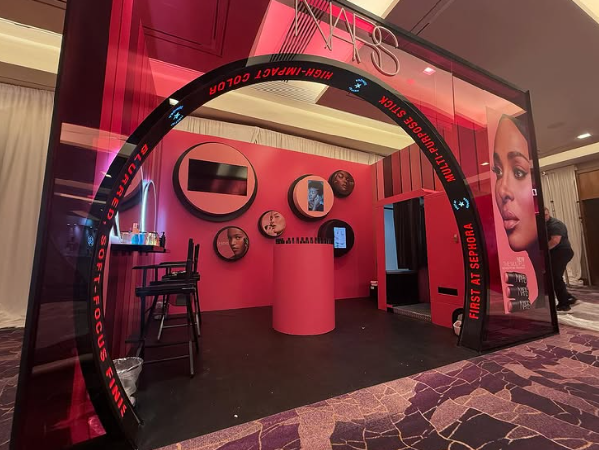 Sephora exhibition booth with pink walls and circular displays of beauty products, featuring a large promotional poster of a woman and makeup items visible on a table.